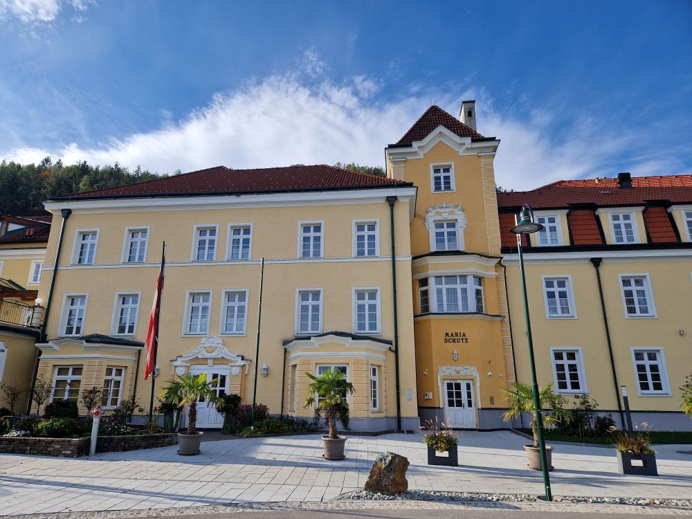 Gelbes Gebäude mit roten Dächern und mehreren Fenstern, umgeben von Pflanzen und Flaggen.