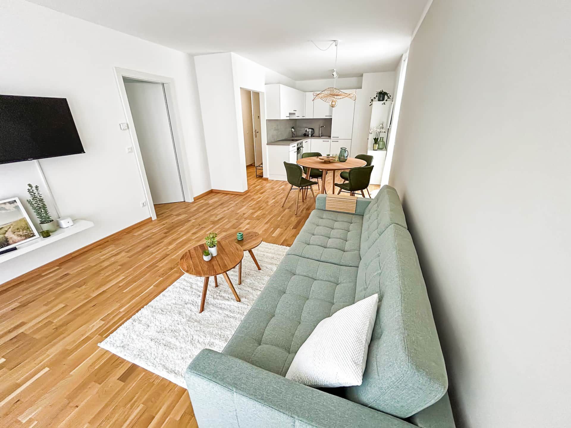 Living room with sage green couch, TV and view into the dining room and kitchen.