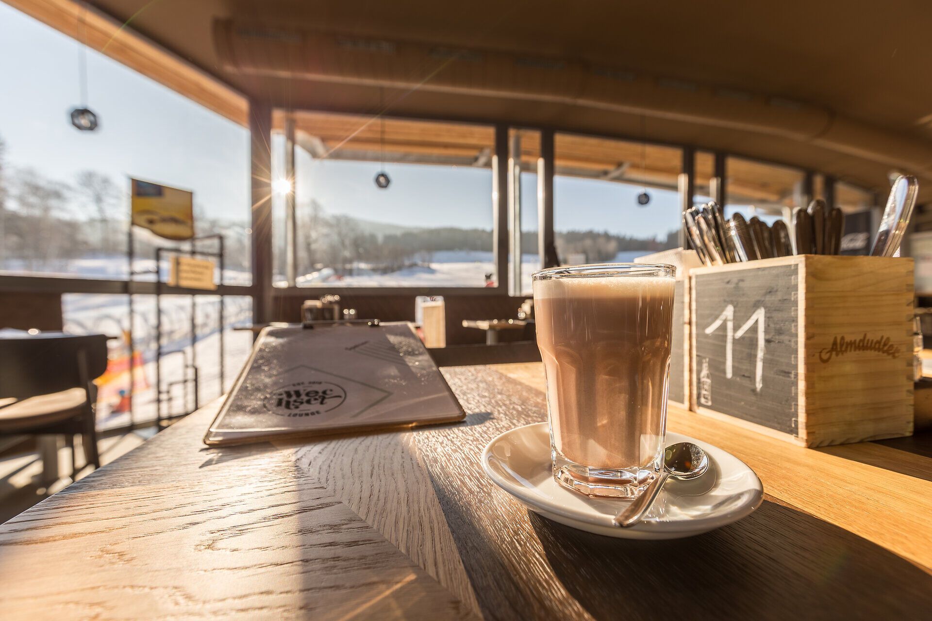 Genießen Sie eine Tasse warmen Kakao in der gemütlichen Atmosphäre eines Berggasthofs, während die Sonne sanft durch die großen Fenster strahlt. Der Blick auf die schneebedeckten Landschaften lädt zum Träumen und Entspannen ein, perfekt für einen unvergesslichen Tag in den Bergen.