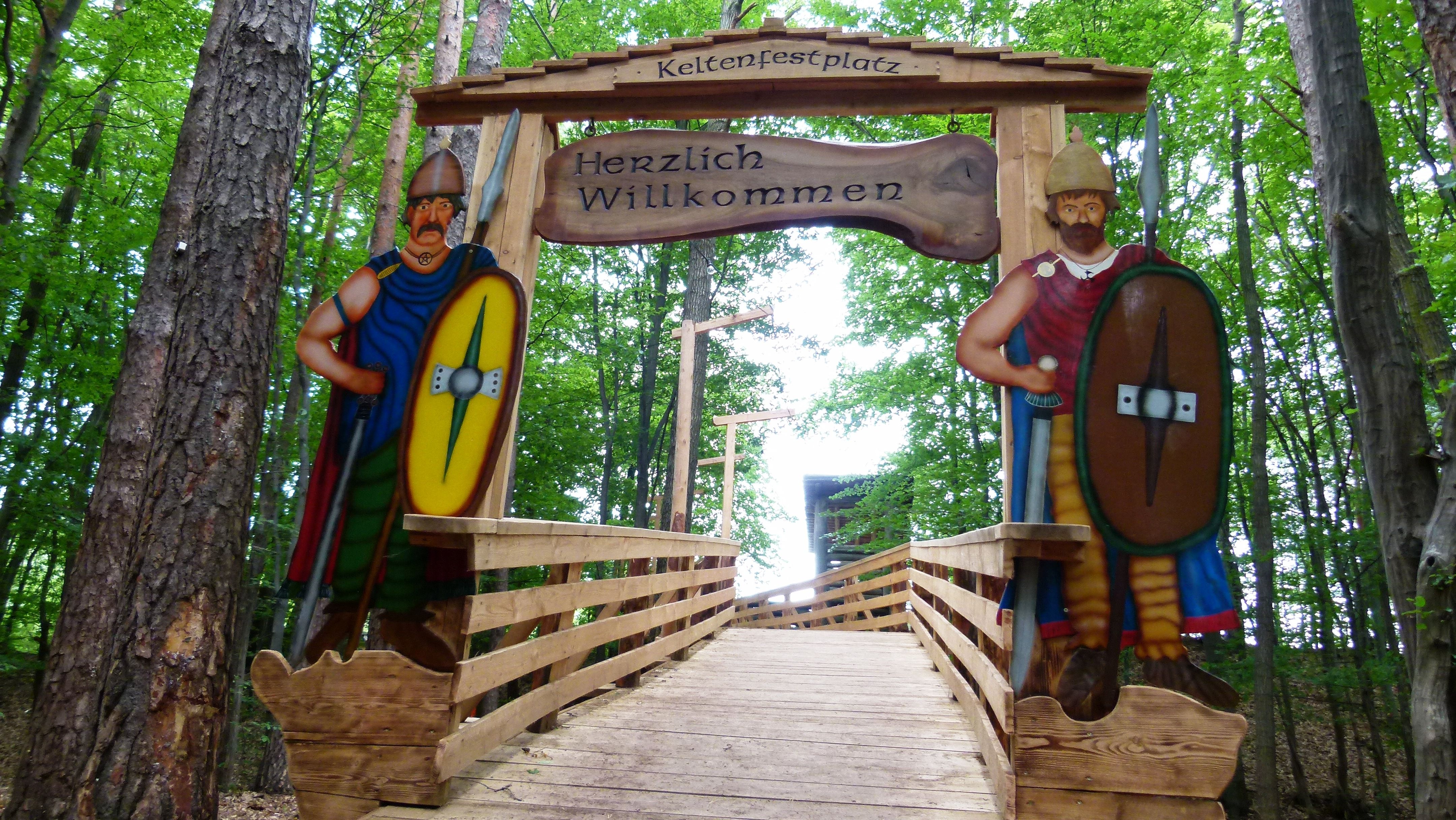 Eine breite Holzbrücke im Wald führt über einen Graben, das Tor zur Holzbrücke ist schön gestaltet und mit Keltenfestplatz und Herzlich Willkommen beschriftet. Links und rechts vom Tor sind 2 Holzplatten montiert, auf denen groß zwei männliche Kelten mit Schild und Speer aufgezeichnet sind.
