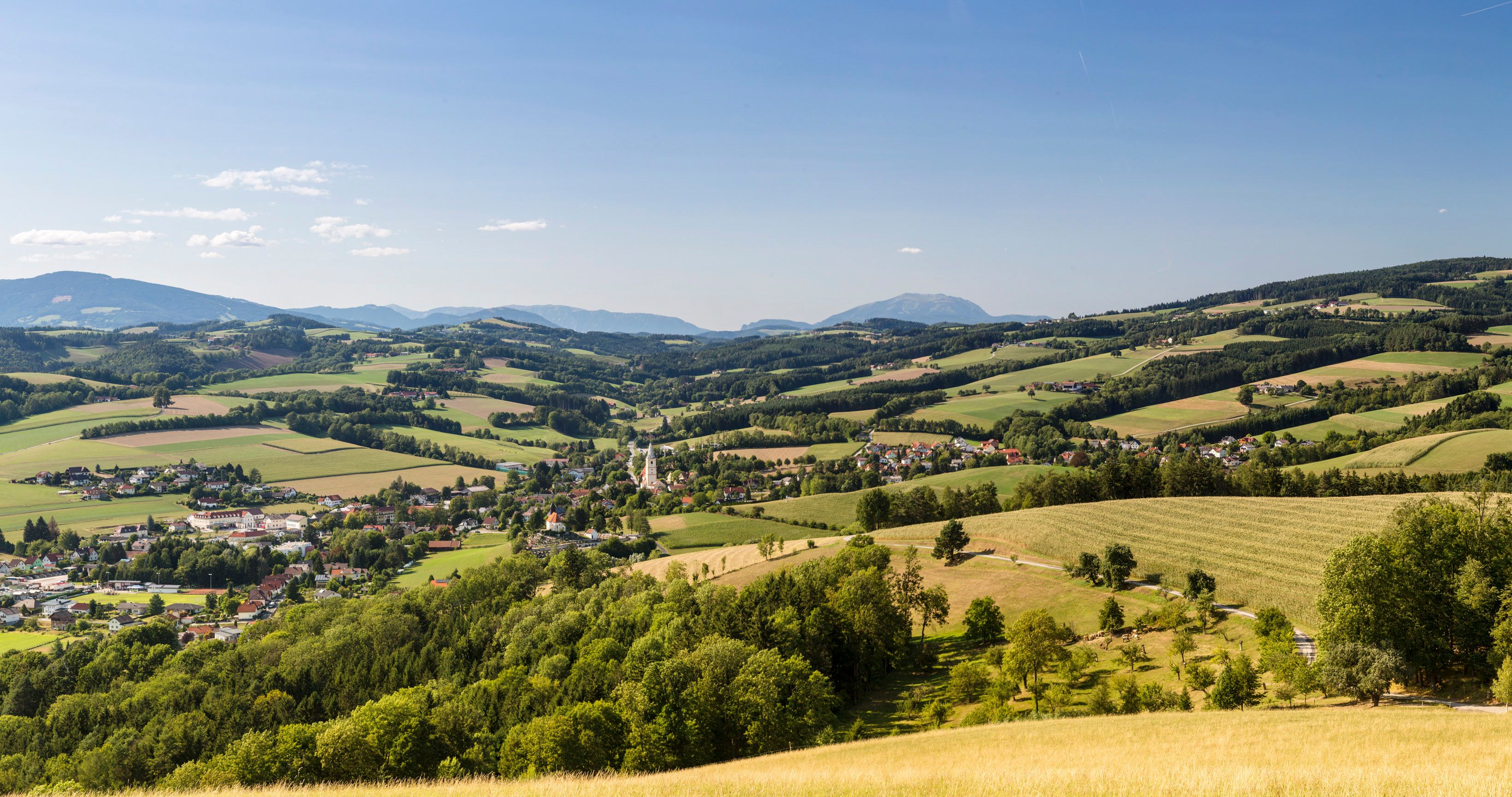 Panoramaaufnahme in der Buckligen Welt, im Tal der Ort Krumbach mit Kirchturm, im Hintergrund Hügel und der Schneeberg.