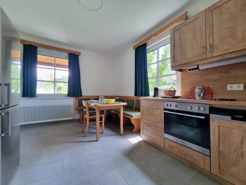 Küche mit Holzmöbeln, Esstisch und Eckbank, zwei Fenstern mit grünen Vorhängen.