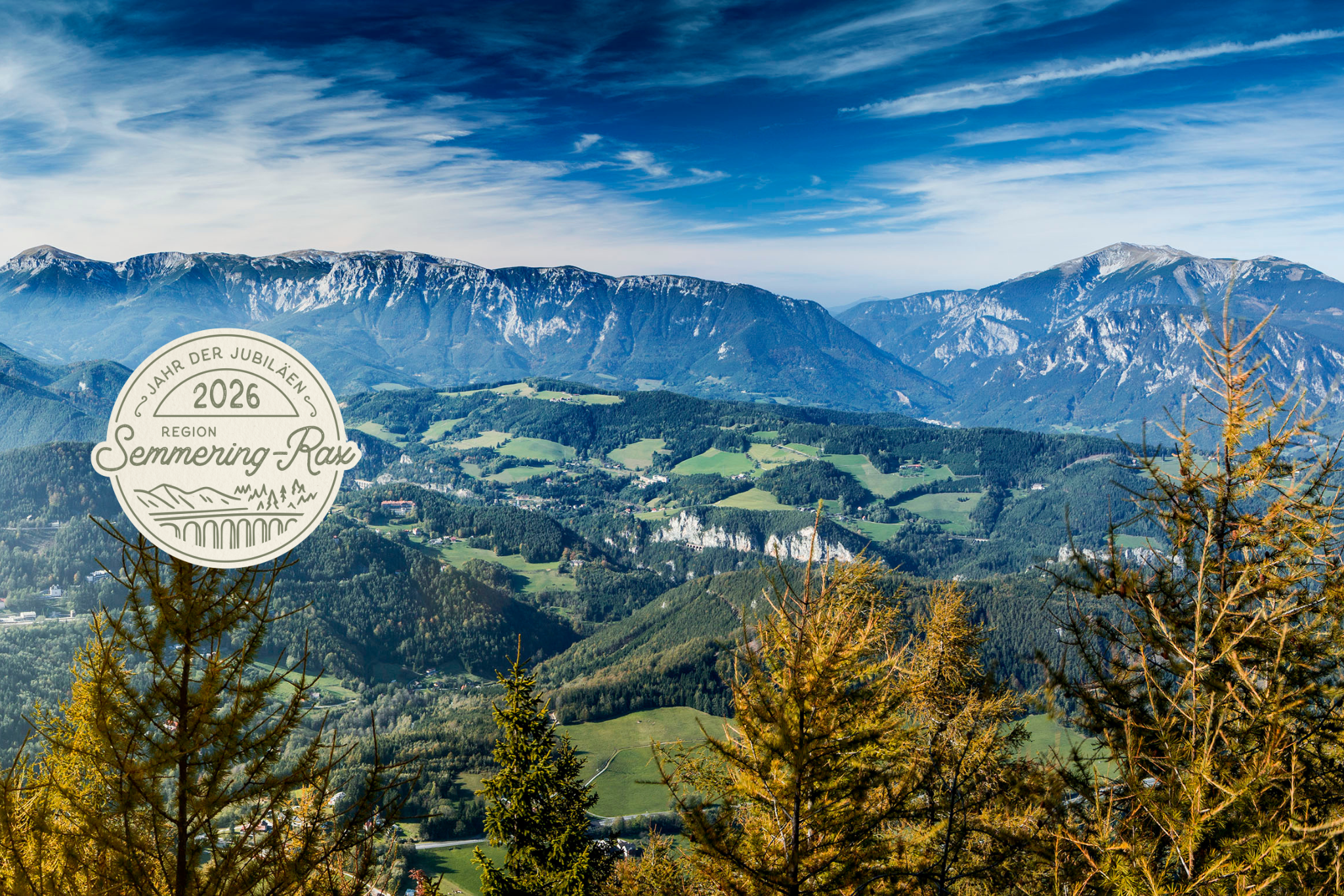 Panoramablick vom Sonnwendstein über die Berglandschaft der Region Semmering-Rax mit grünen Tälern, bewaldeten Hängen und markanten Felswänden unter blauem Himmel, inklusive Button zum Jubiläumsjahr 2026.