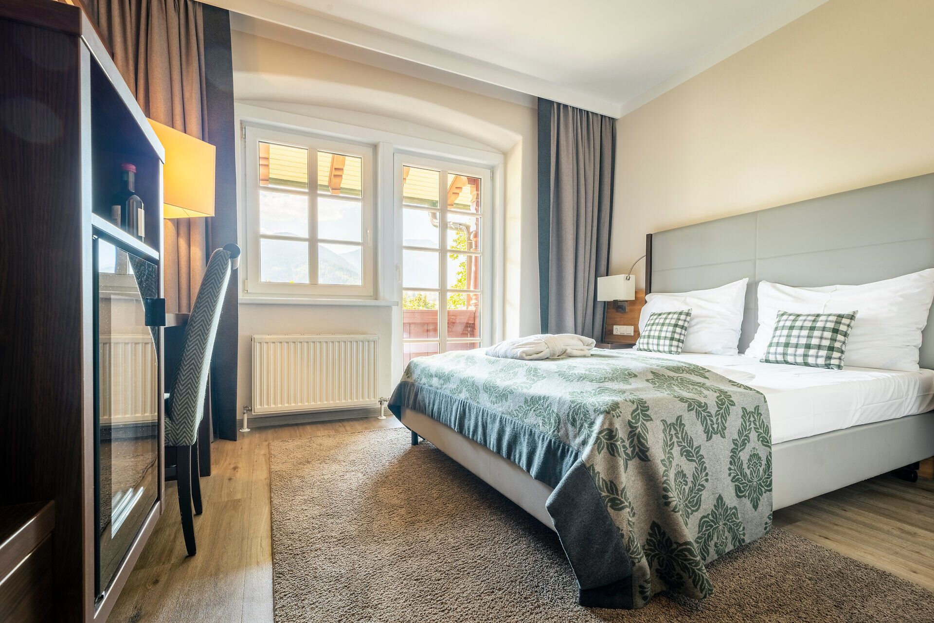 Im gemütlichen Zimmer vom Hotel Schneeberghof mit Fenster und Balkontüre befindet sich ein großes Doppelbett, gegenüber ein Schrank und ein Tisch mit Stuhl.