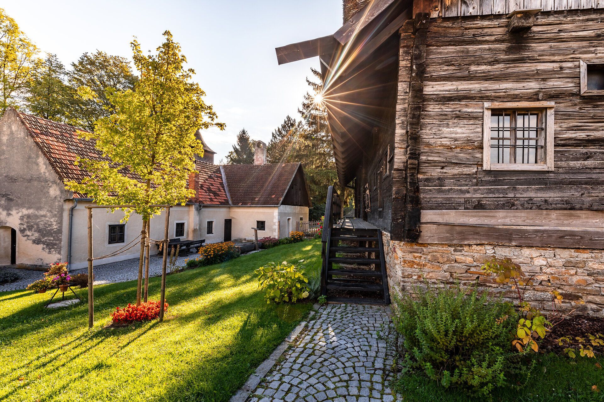 Blick auf ein traditionelles Holzhaus mit Terrasse und auf ein gemauertes Haus sowie den Garten im Museumsdorf Krumbach bei Sonnenschein 