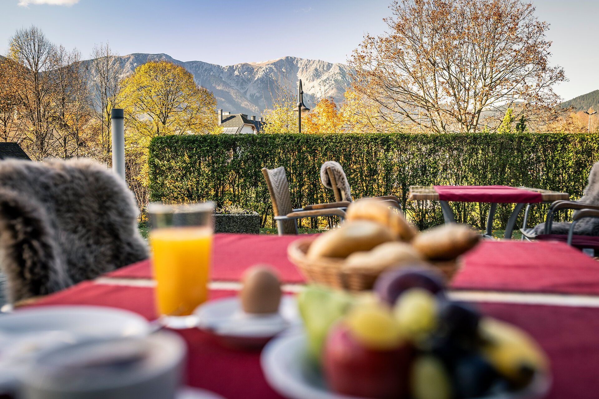 Auf der Terrasse vom Hotel Schneebergbof stehen vor einer gepflegten Hecke und Bergaussicht zwei Tische mit Sesseln und roten Tischtüchern, ein Tisch ist für das Frühstück gedeckt. 