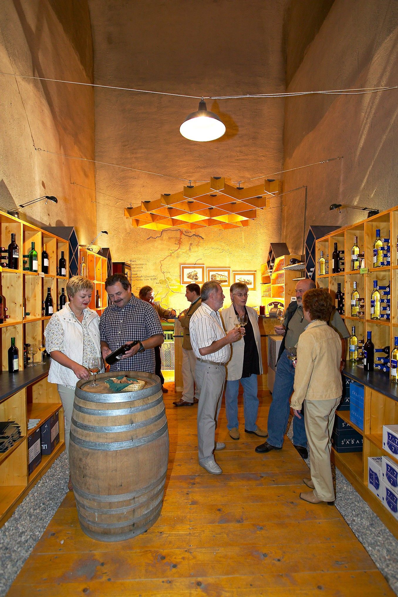 Menschen in einem Weinkeller oder Vinodukt mit Holzregalen, Weinflaschen und einem Fass im Vordergrund, sie probieren Wein und unterhalten sich.