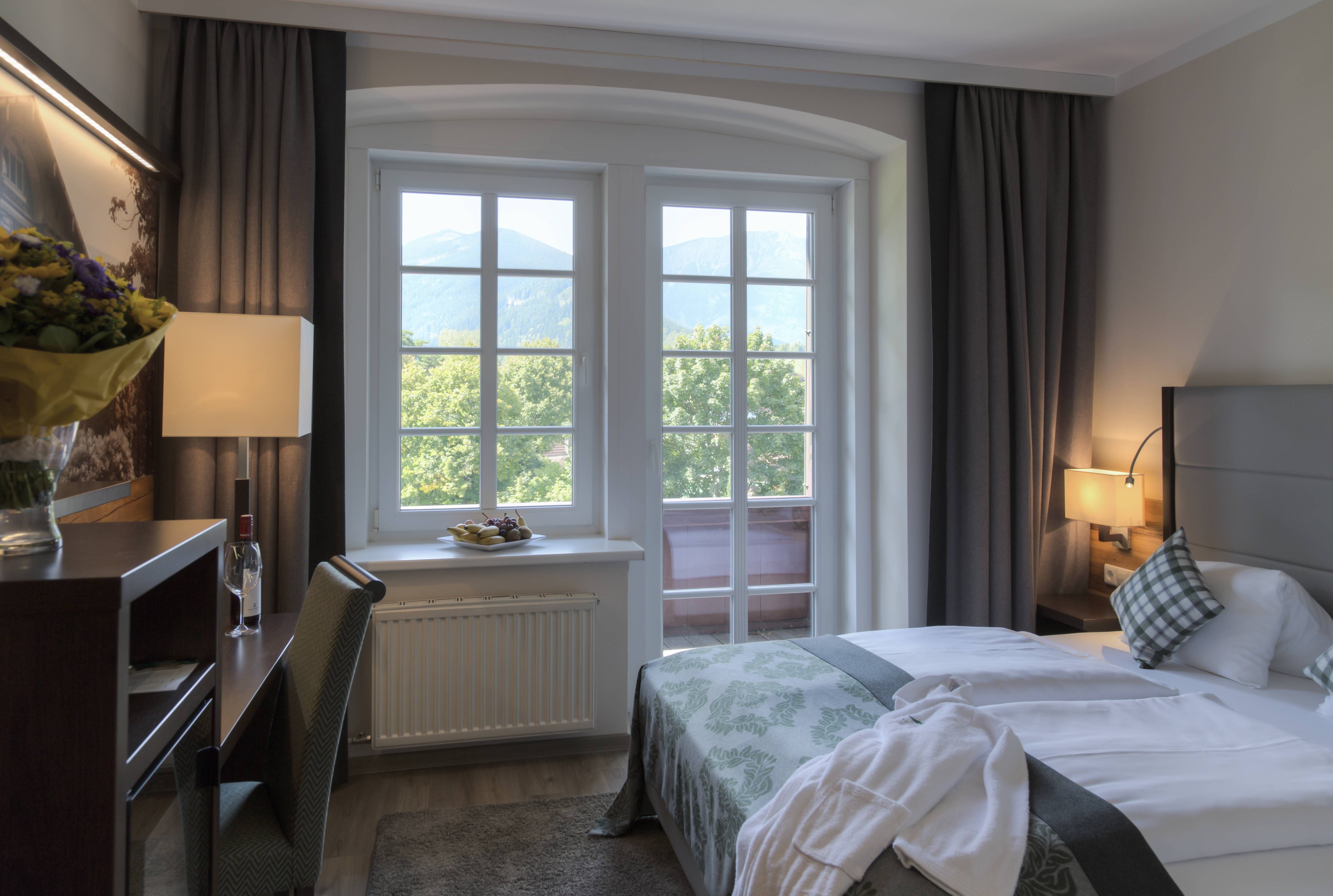 Ein gemütliches Doppelzimmer mit einem großen Fenster, das einen Blick auf die Berge bietet. Das Zimmer ist mit einem Bett, einem Schreibtisch und einer Lampe ausgestattet.