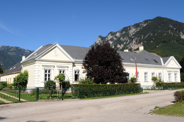 Ein großes, helles Gebäude mit Garten vor einer Bergkulisse.