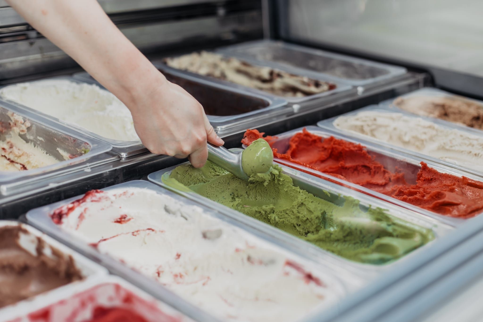 Eine Hand schöpft mit einem Eisportionierer Eis aus einer Vitrine mit diversen Eissorten.