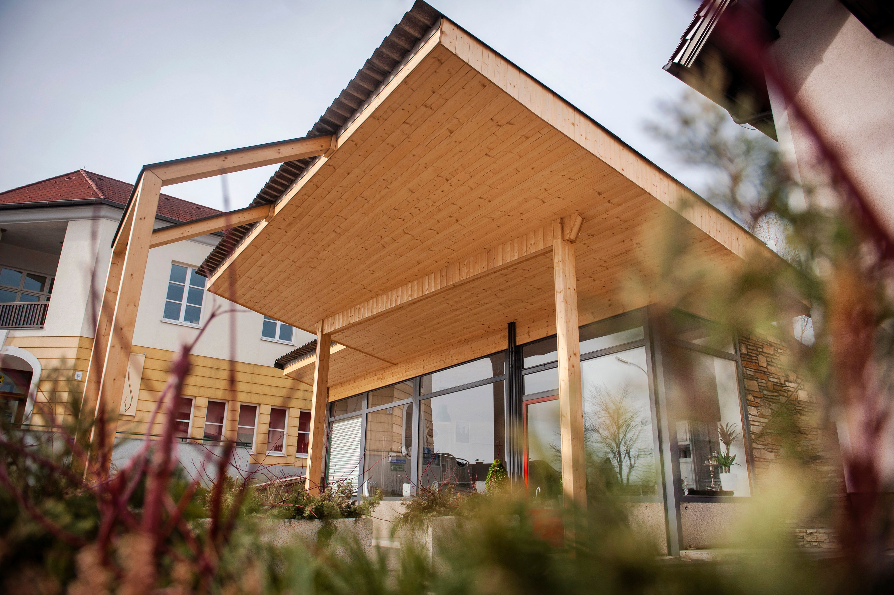 Modernes Gebäude mit Holzüberdachung und Glasfront, umgeben von Pflanzen.