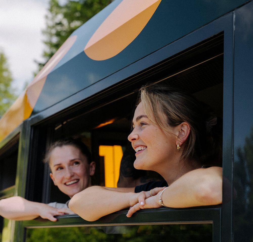 Two cheerful women look out the window of the Schneebergbahn's Salamander train as the wooded landscape passes by in the background, reflecting their joy and curiosity about the journey ahead.