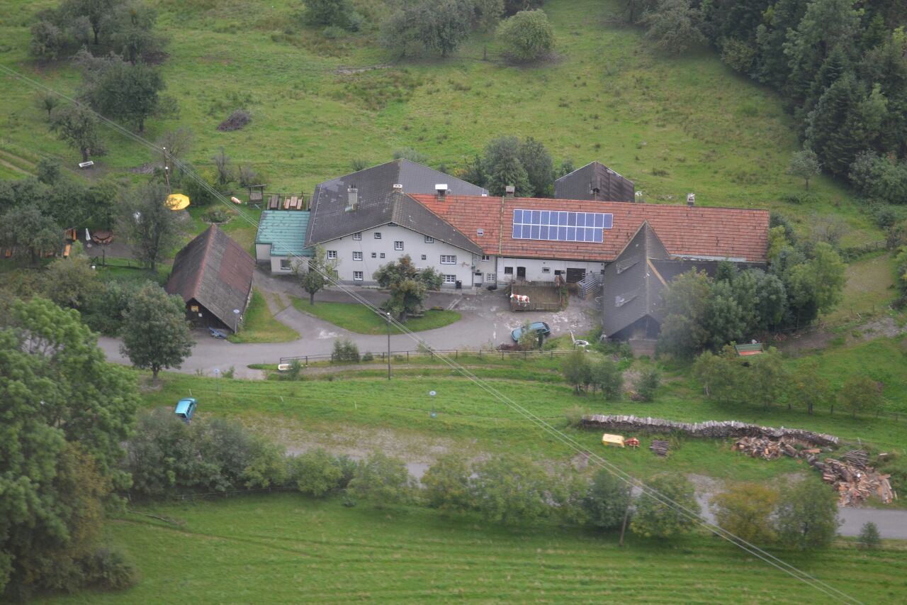 Luftaufnahme eines Bauernhofs mit Solarpanelen auf dem Dach, umgeben von grünen Feldern und Bäumen.