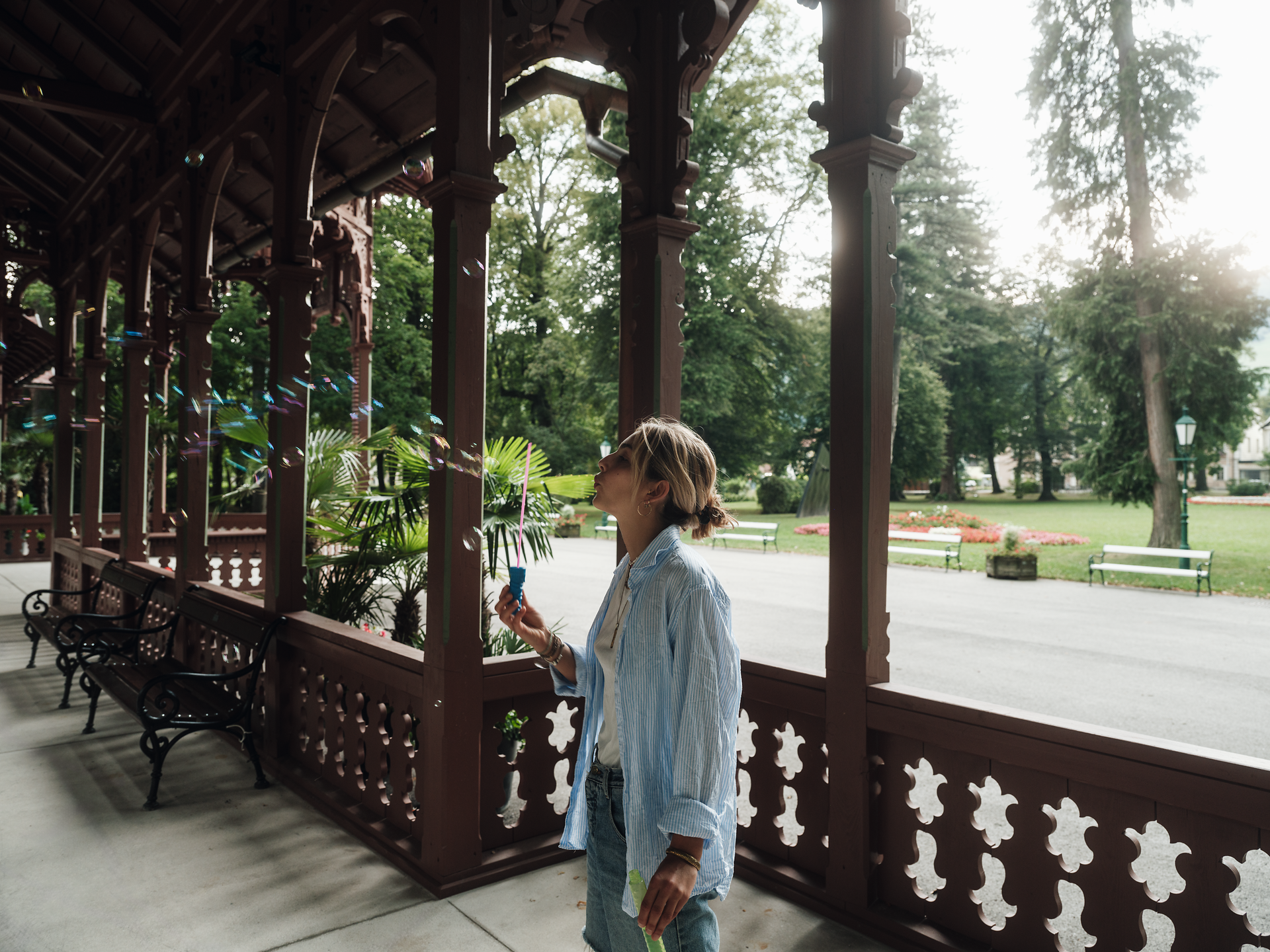 Ein sanfter Sommerwind weht durch die malerische Landschaft, während die Sonne sanft auf die bunten Blumen im Garten scheint. Eine entspannte Atmosphäre umgibt die Szene, in der eine Person fröhlich Seifenblasen in die Luft bläst und die Schönheit der Natur genießt.