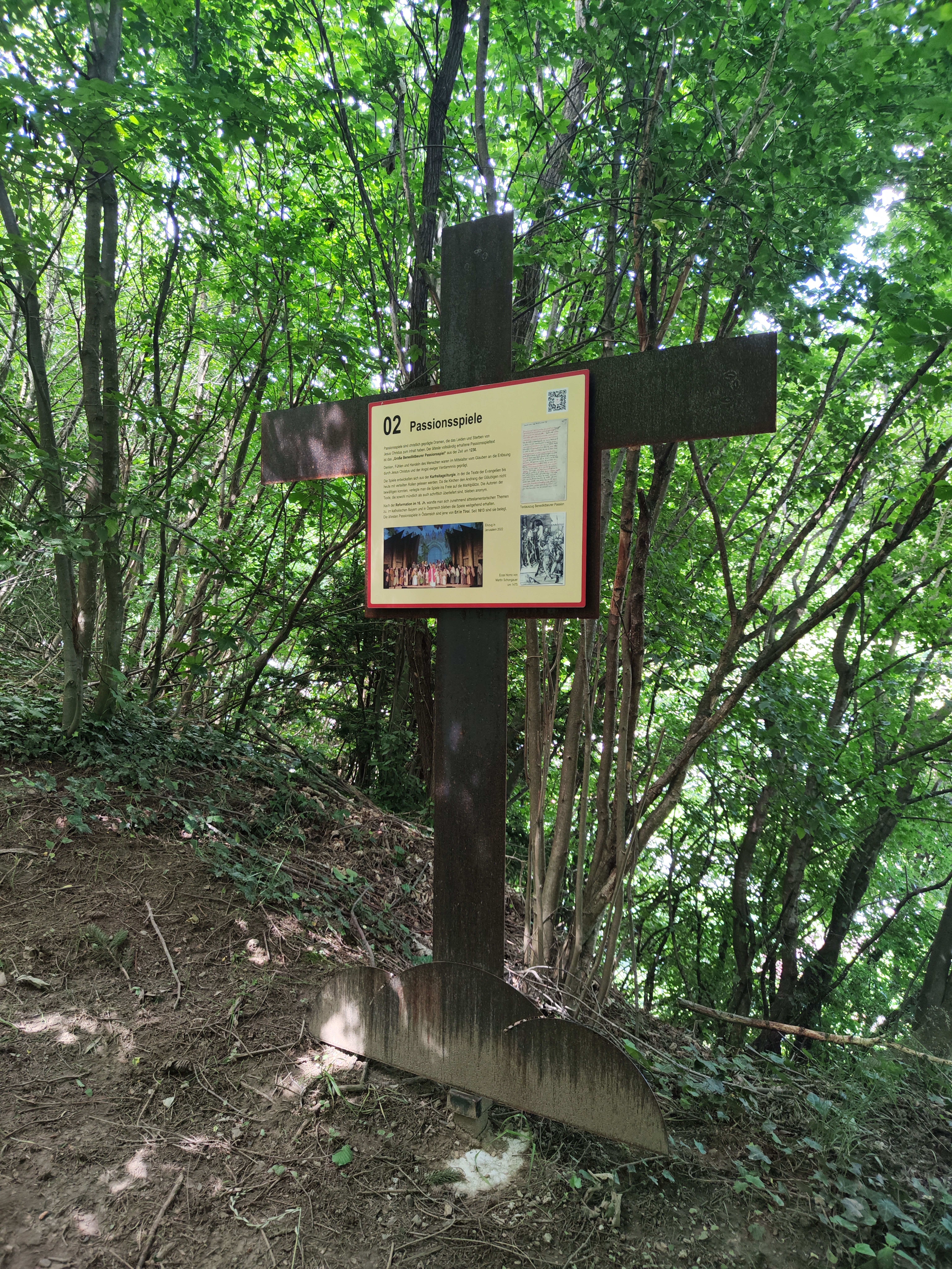 Ein Schild über die Passionsspiele an einem Kreuz auf einem Waldweg.
