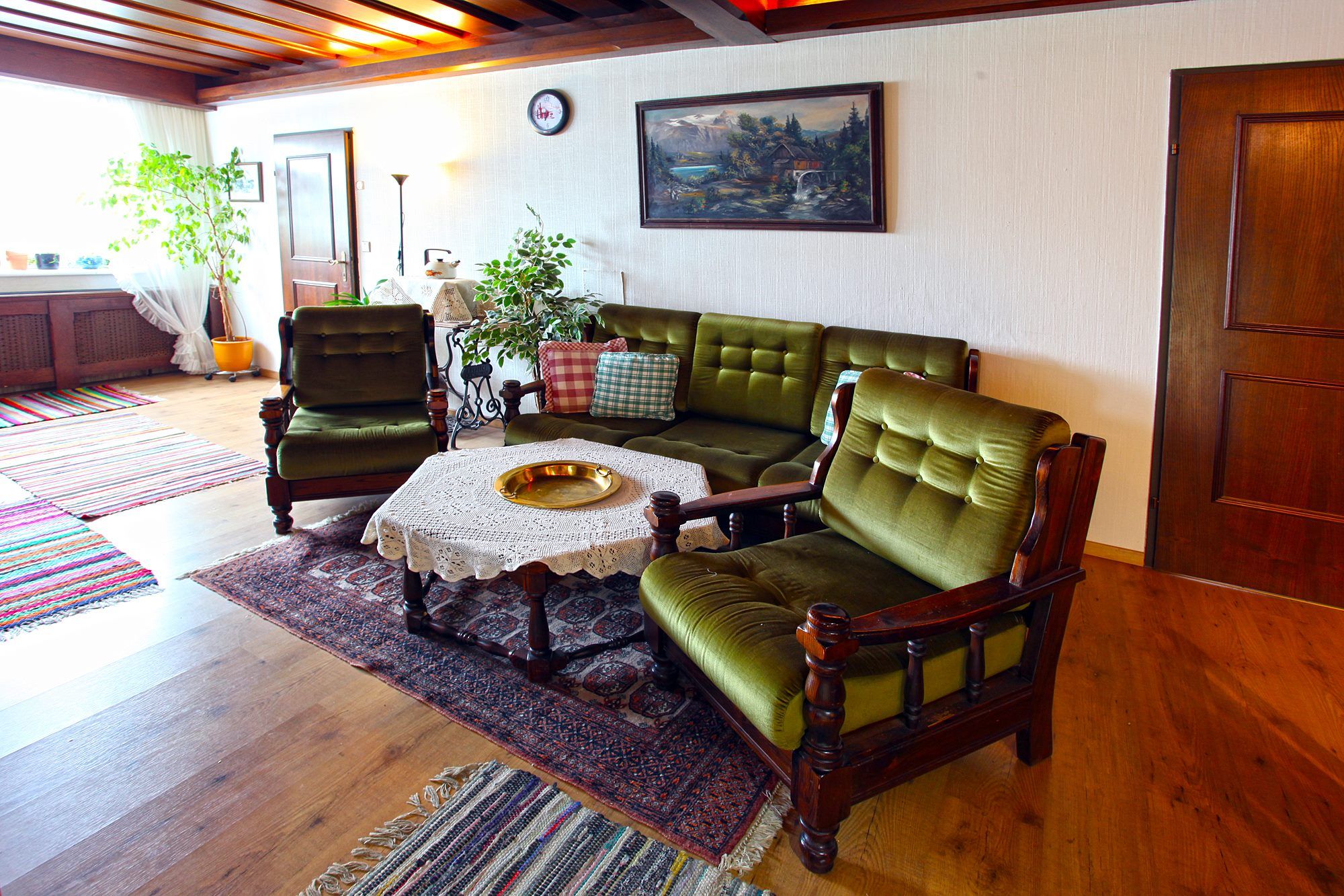 Wohnzimmer mit grünen Polstermöbeln, Holztisch mit Spitzendecke, Pflanzen und Wandbild.