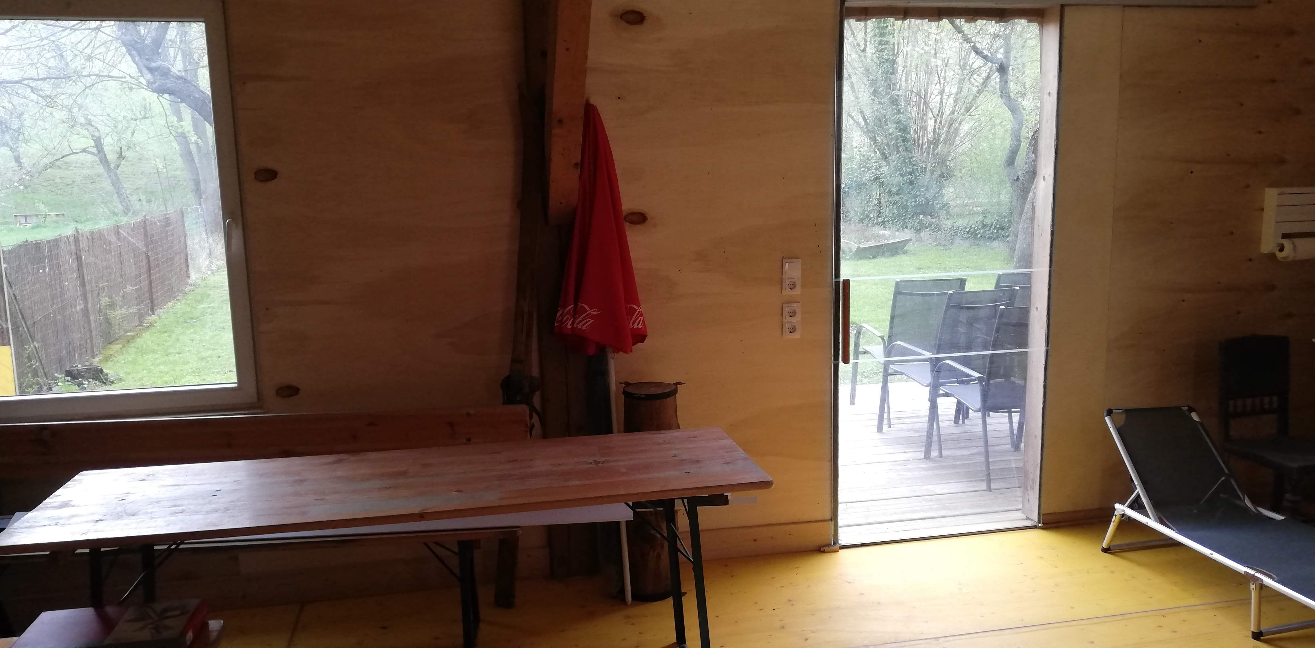 Innenansicht eines Raumes mit Holzwänden, Tisch, Liegestuhl und Blick auf Terrasse und Garten.