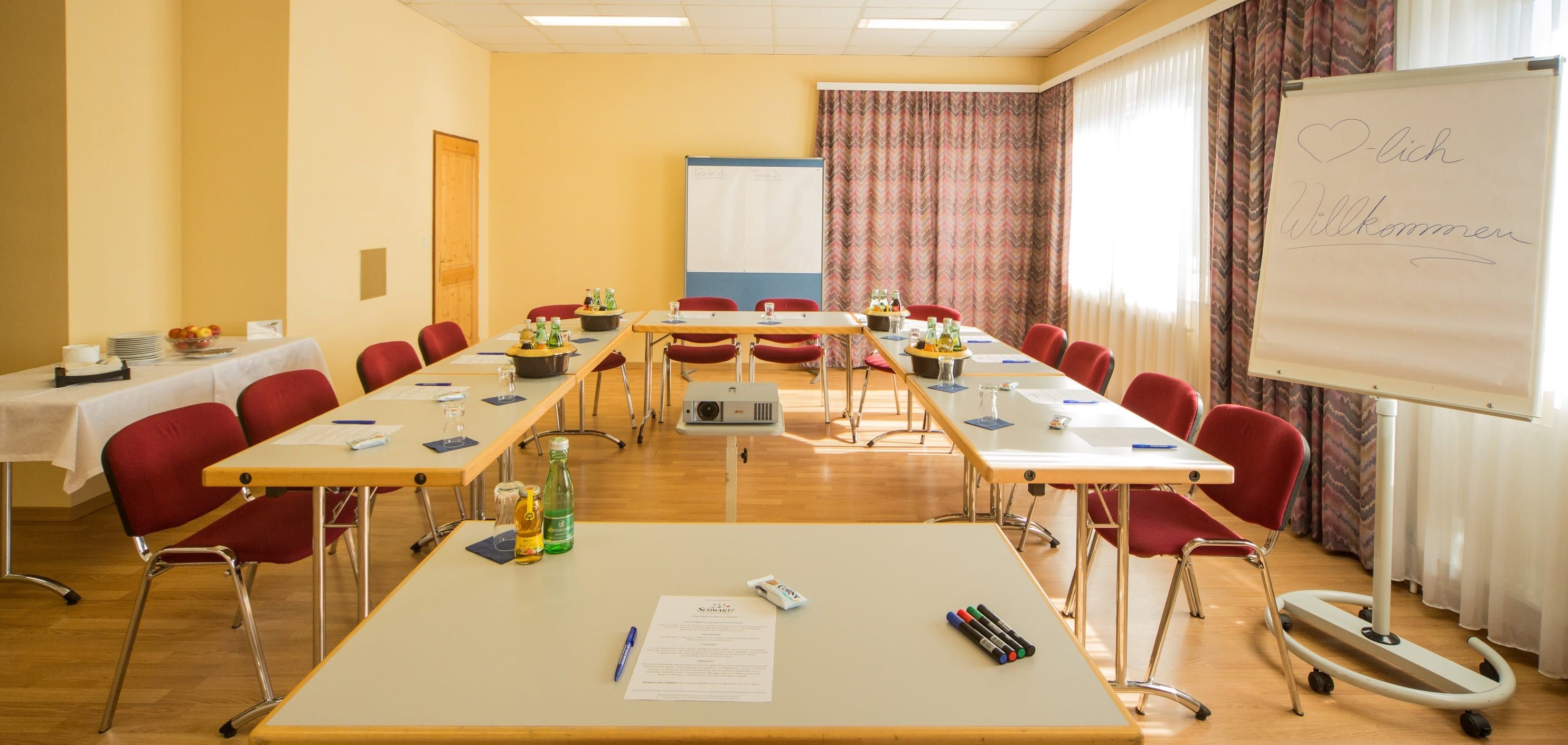 Seminarraum mit U-förmiger Tischanordnung, Flipchart und Getränken.