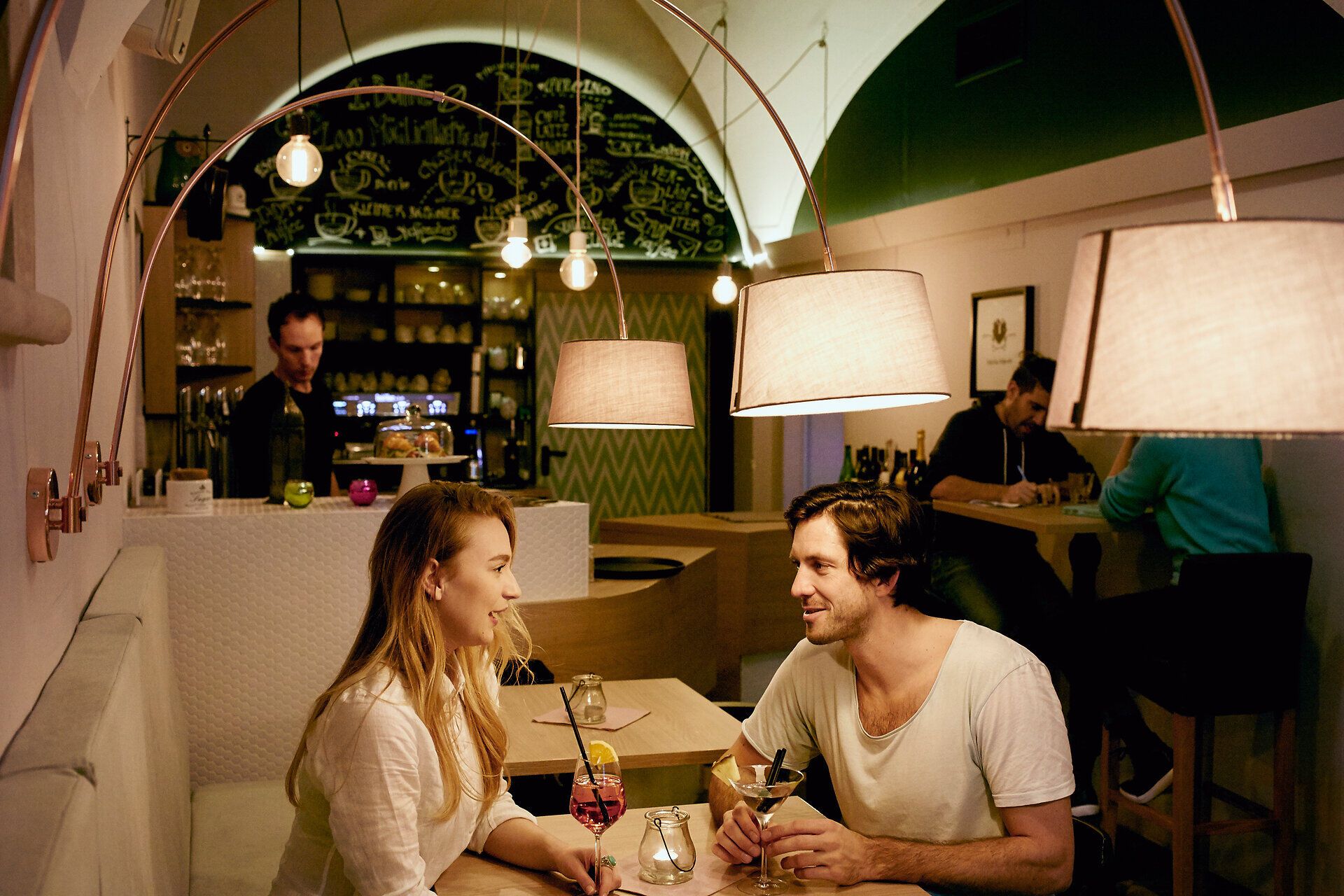 Ein Pärchen sitzt auf einem Tisch in einem Lokal mit Weinglas und Martiniglas und stoßen an.