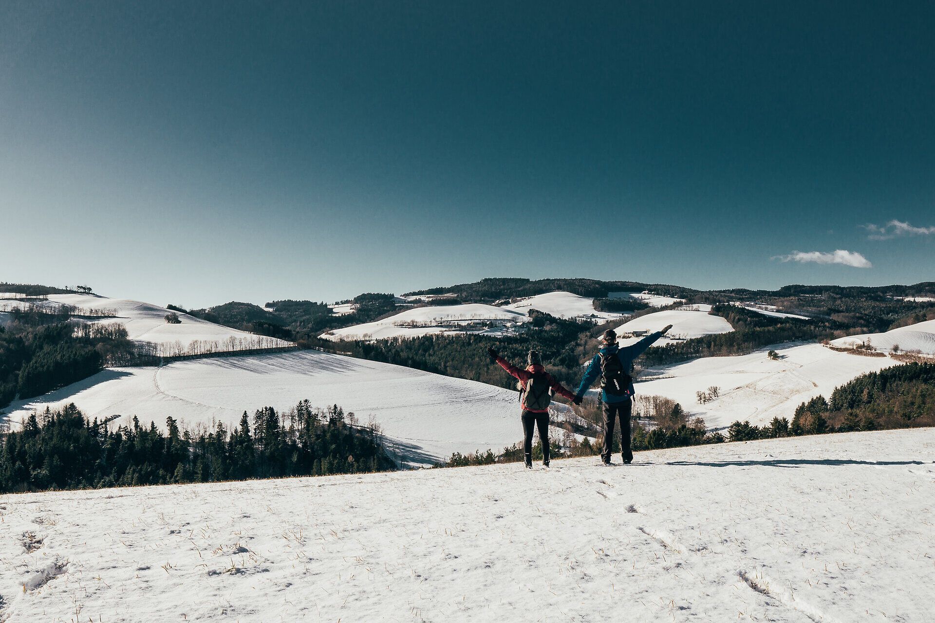 In der winterlichen Landschaft erheben sich die schneebedeckten Hügel majestätisch gegen den klaren blauen Himmel. Zwei Wanderer genießen die frische Luft und die atemberaubende Aussicht, während sie den Weg der Blicke entlang schreiten. Die Stille der Natur und die glitzernde Schneedecke schaffen eine friedliche Atmosphäre, die zum Verweilen einlädt.