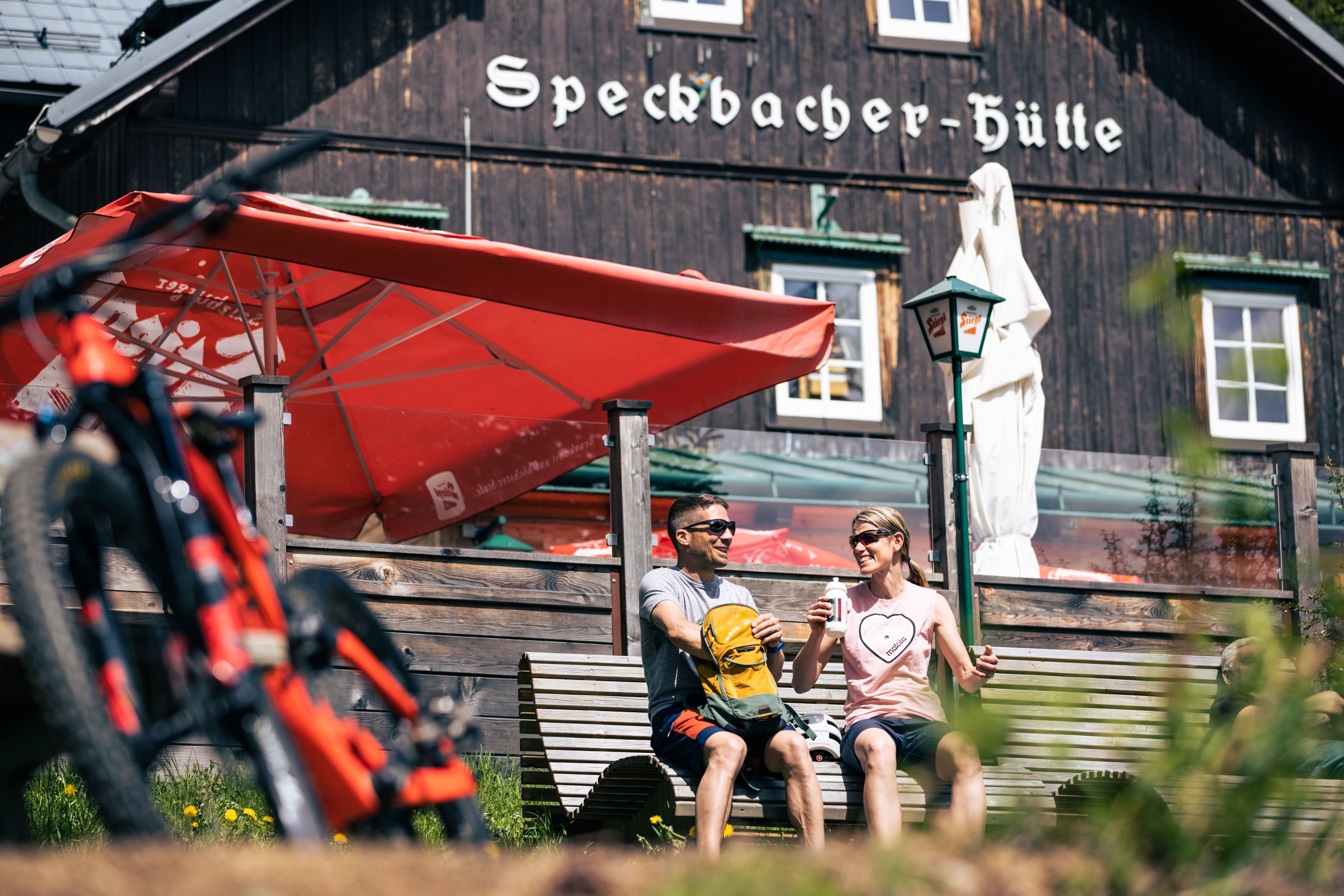 Zwei Mountainbiker sitzen auf einer Bank vor der Speckbacherhütte, seitlich sieht man die Räder.