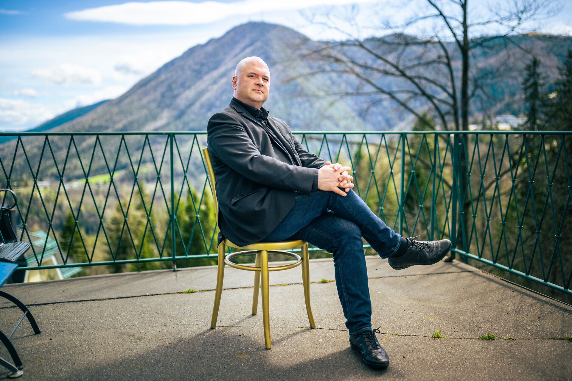Mann im dunklen Sakko sitzt auf einer Terrasse mit Blick auf Berge und Tallandschaft.