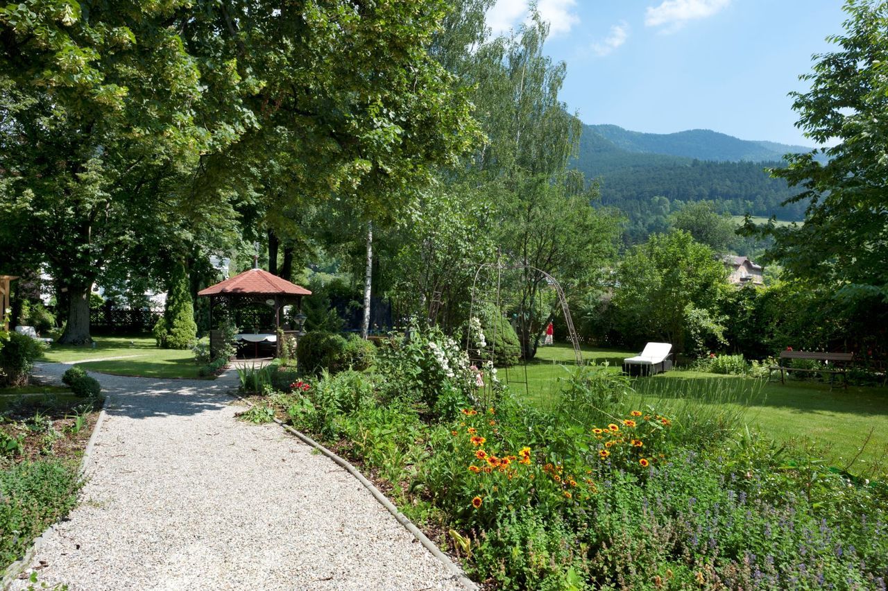Ein gepflegter Garten mit Kiesweg, Blumenbeeten, einem Pavillon und einer Liege. Im Hintergrund sind Bäume und Berge zu sehen.