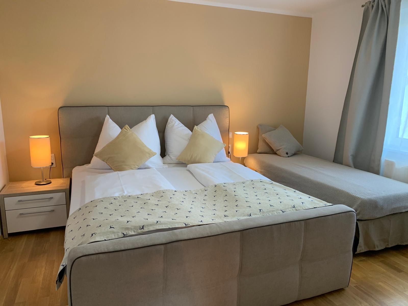 Gemütliches Schlafzimmer mit Doppelbett und Einzelbett, zwei Nachttischlampen und Vorhängen.