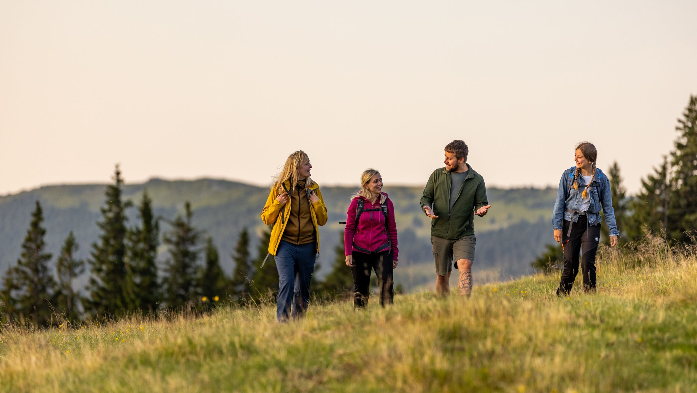 Vier Personen wandern auf einer Wiese in den Bergen bei Sonnenuntergang.
