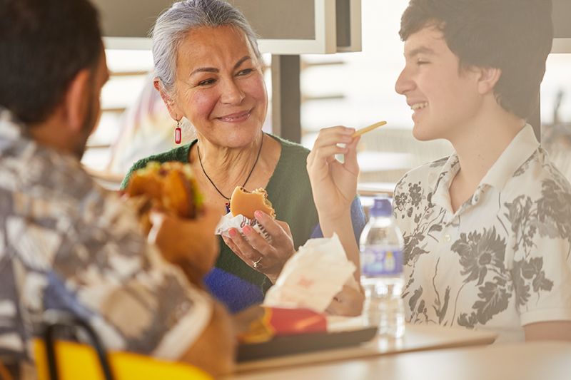 Drei Personen sitzen an einem Tisch und essen Fast Food. Eine ältere Frau lächelt einen jungen Mann an, der eine Pommes in der Hand hält.