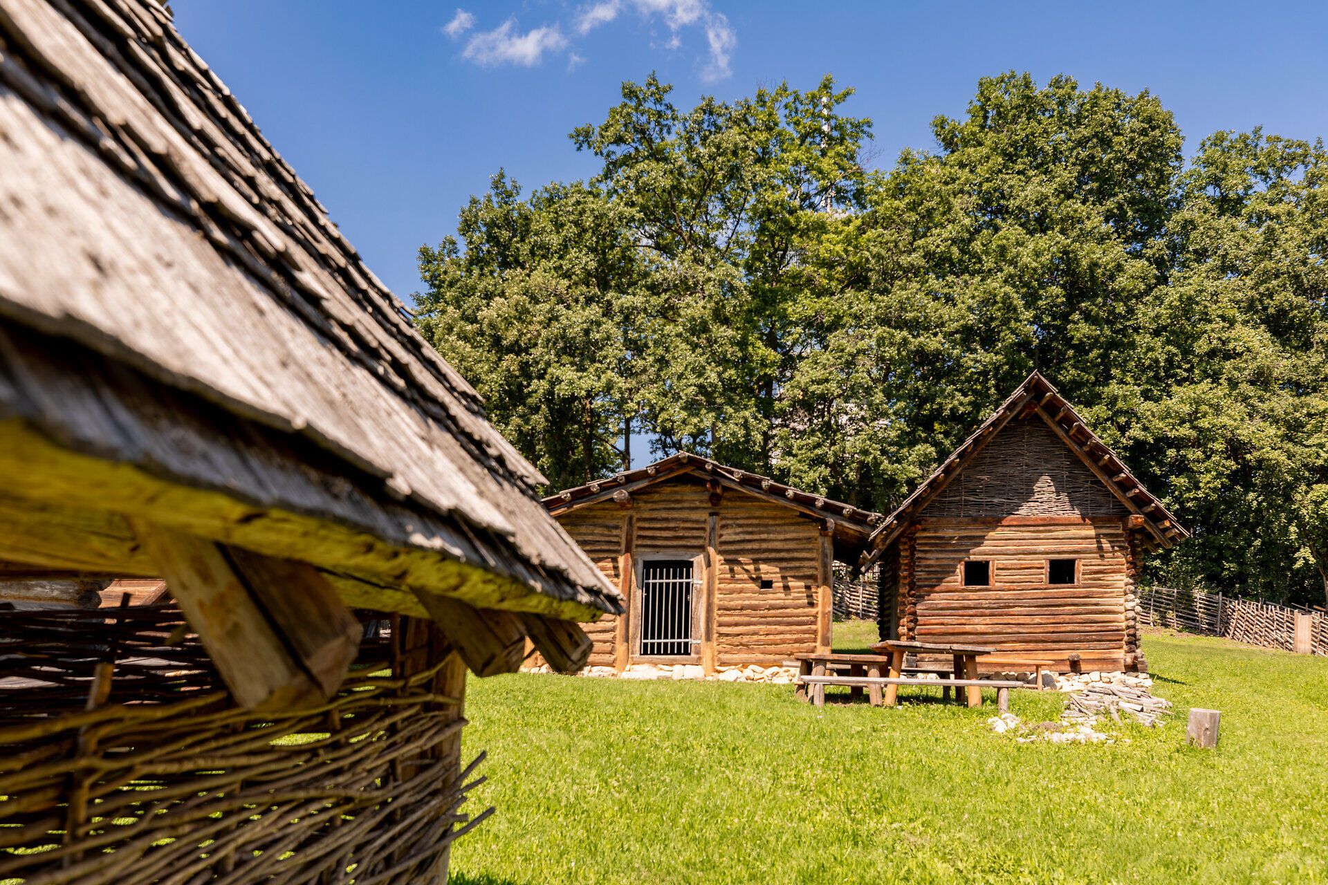 Außenansicht von drei Holzhäusern im Keltendorf Schwarzenbach. Das linke Haus im Vordergrund ist angeschnitten, die beiden anderen Häuser stehen weiter hinten in der Wiese.