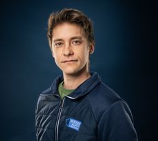 Portraitfoto Wiener Alpen Mitarbeiter, der eine Jacke mit Wiener Alpen Logo trägt und vor einem dunklem Hintergrund steht.