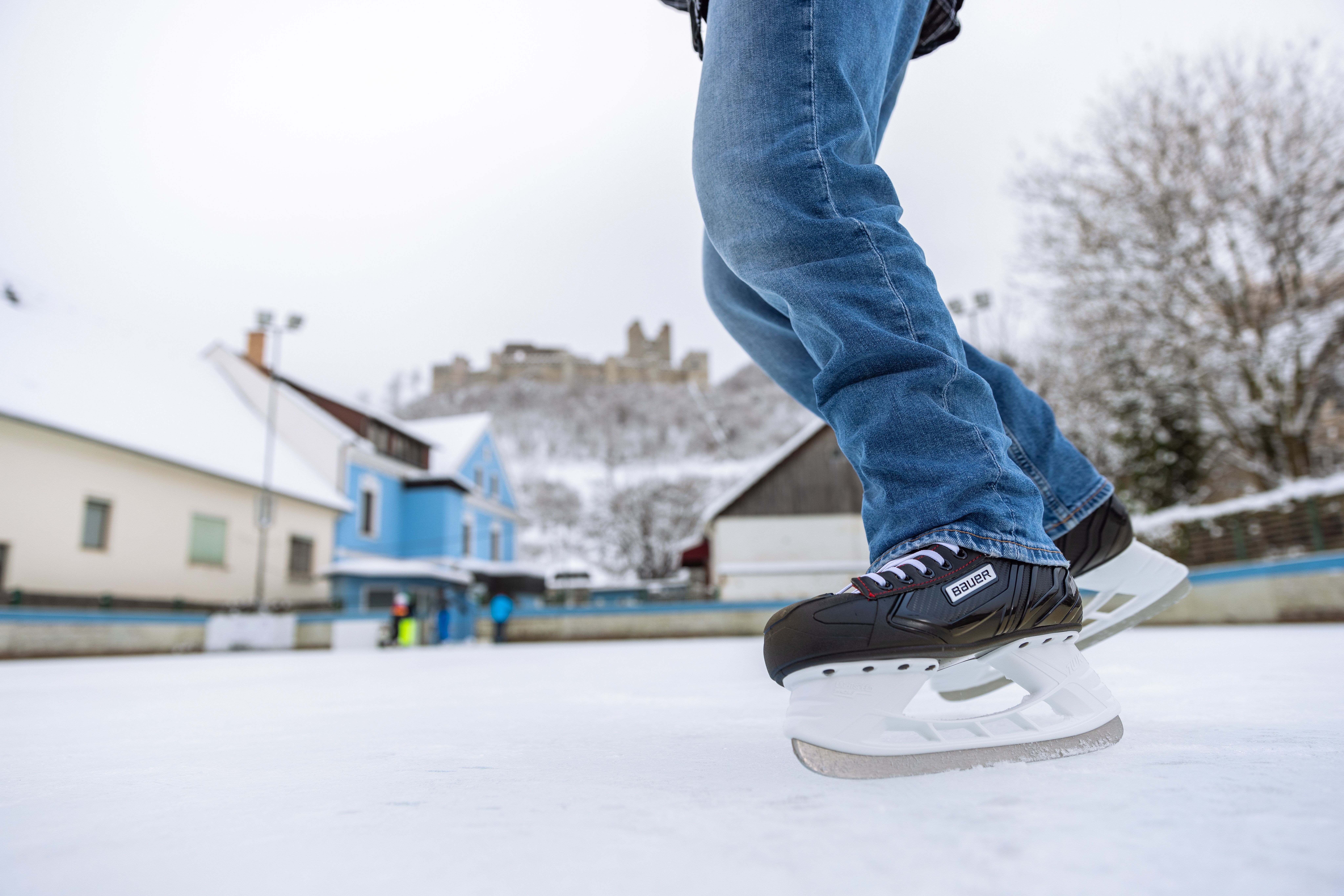 Nahaufnahme von Beinen in Schlittschuhen, die über den Eislaufplatz fahren, im Hintergrund die Burgruine Kirchschlag auf einem bewaldeten Hügel