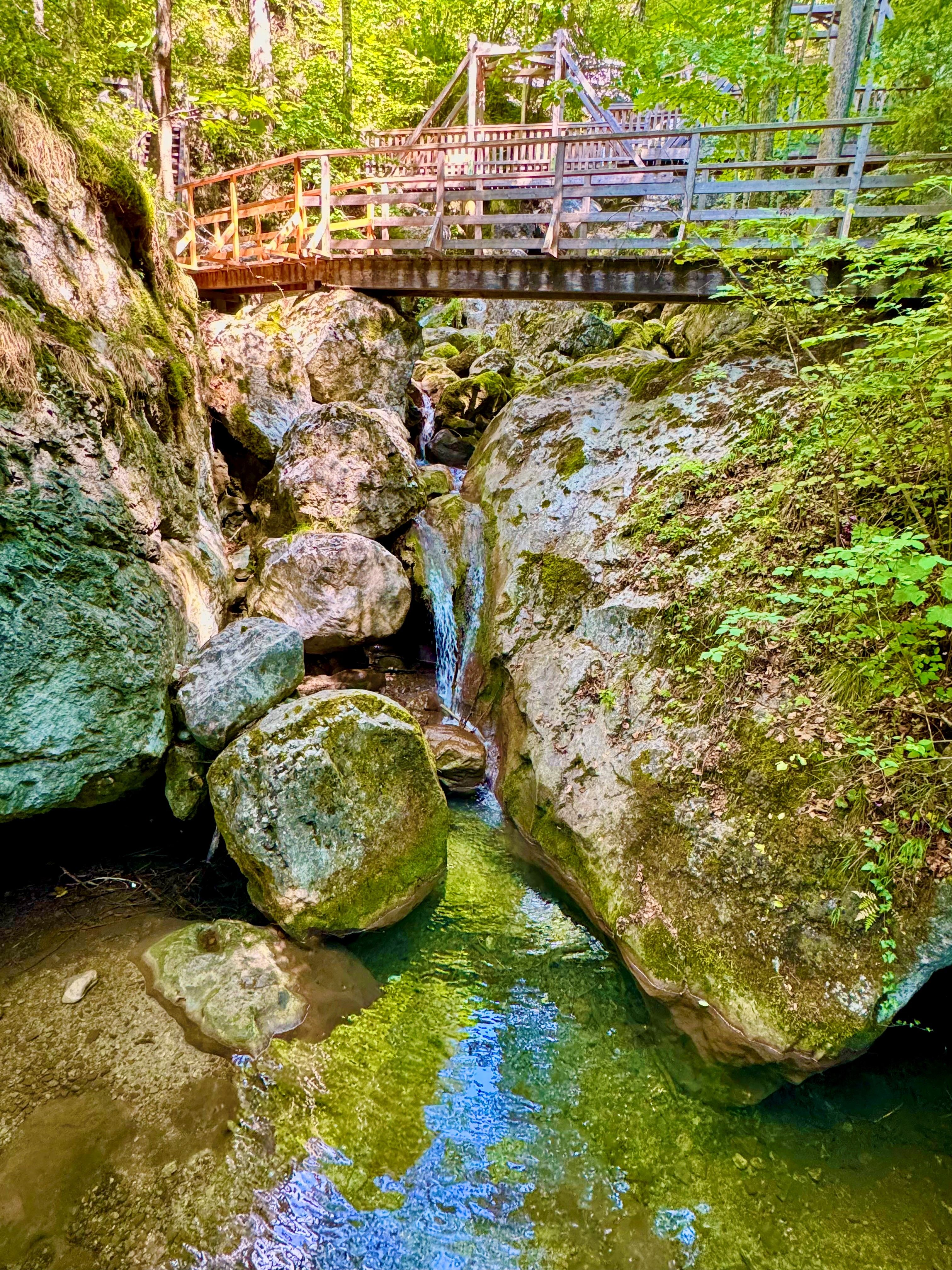 Ein kleiner Wasserfall fließt zwischen großen Felsen unter einer Holzbrücke hindurch, umgeben von üppigem Grün.