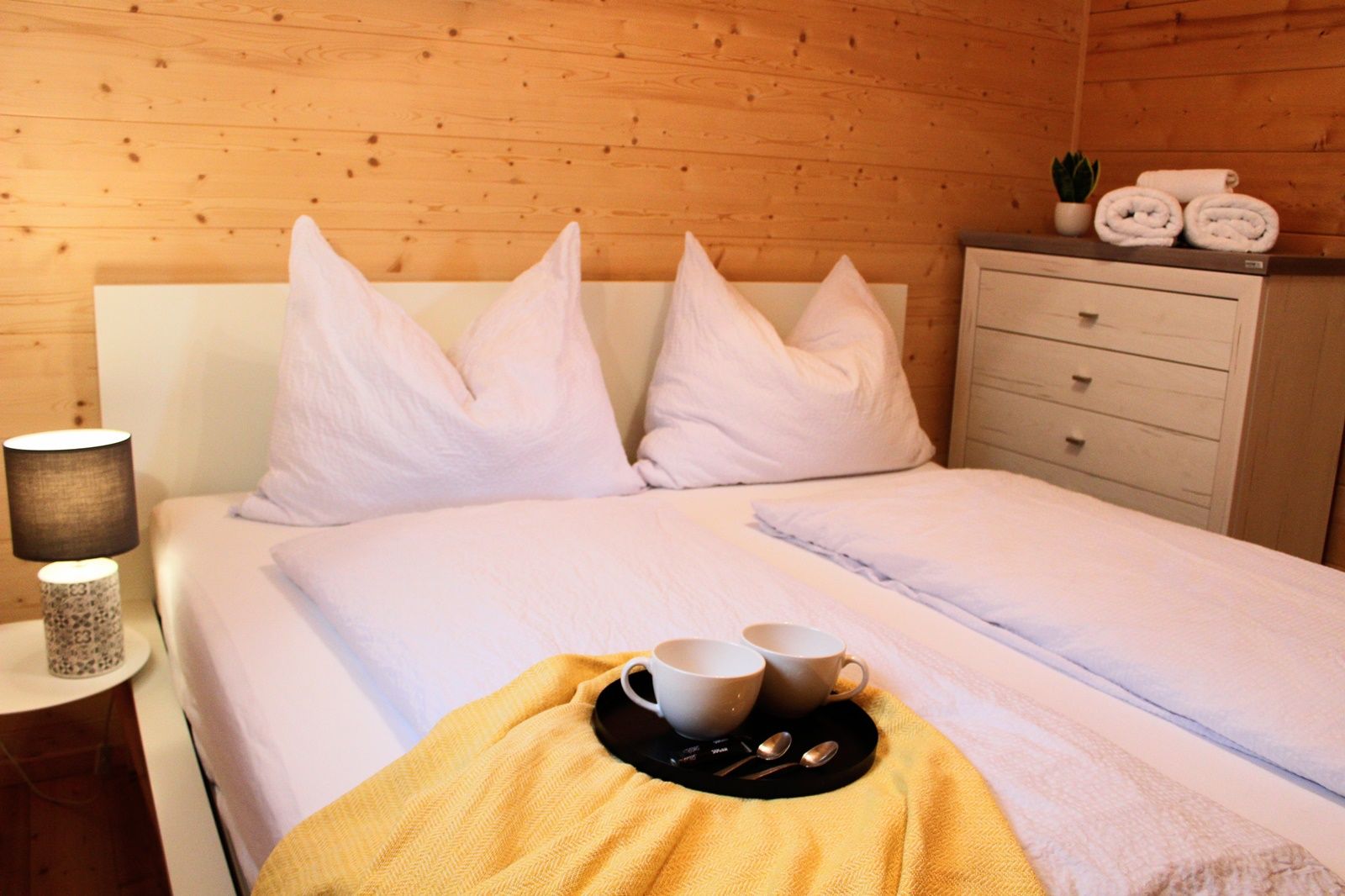 Gemütliches Schlafzimmer mit Doppelbett, weißen Kissen und Decken, gelber Decke, Tablett mit Tassen, Nachttischlampe und Kommode mit Handtüchern.
