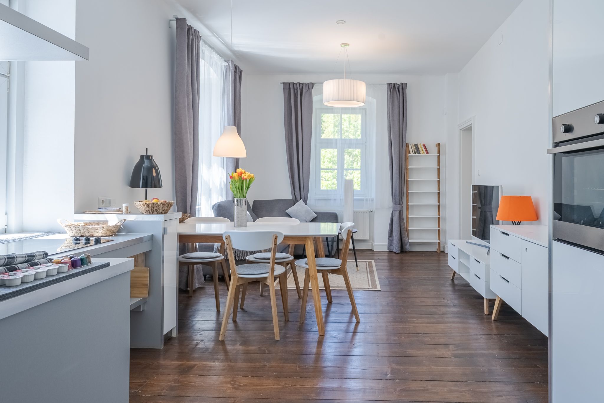 Helles, modern eingerichtetes Wohnzimmer mit Holzboden, Esstisch, Stühlen und Sofa. Große Fenster mit grauen Vorhängen lassen viel Licht herein.