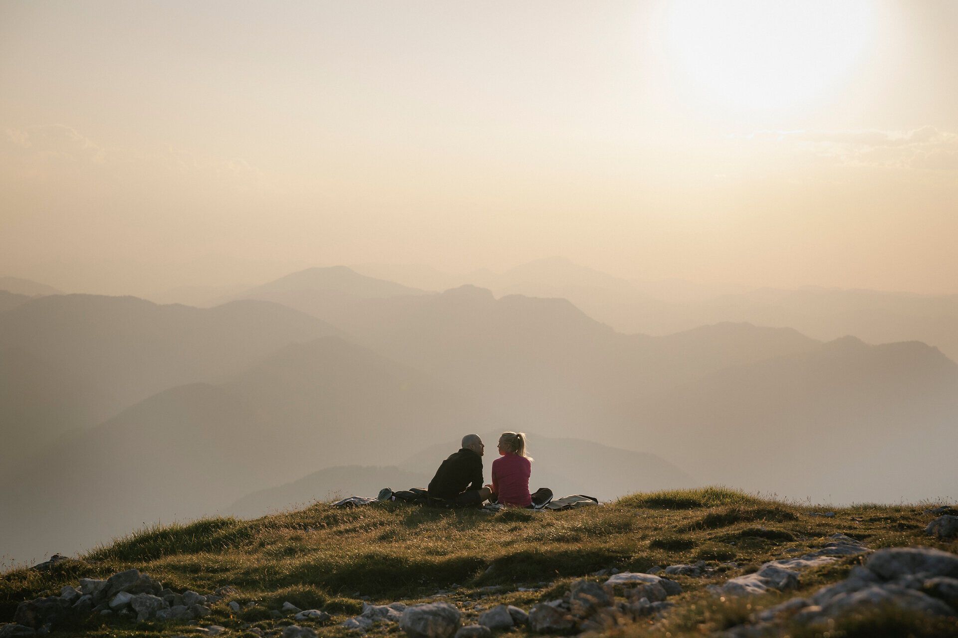 Zwei Personen sitzen im Abendlicht auf einer Wiese am Schneeberg und blicken über die Berglandschaft