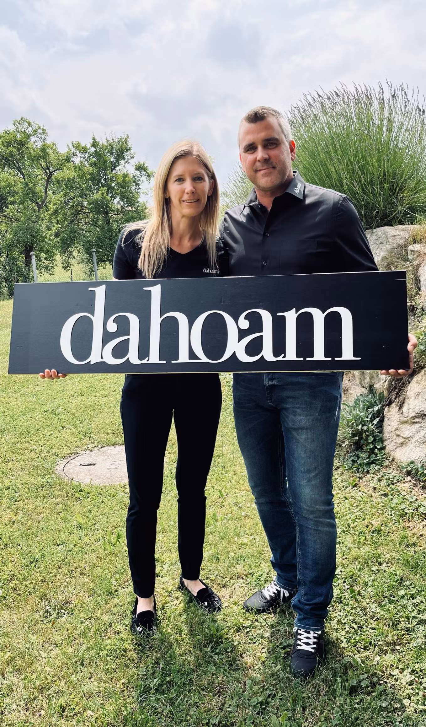 Zwei Personen halten ein Schild mit der Aufschrift 'dahoam' im Freien.