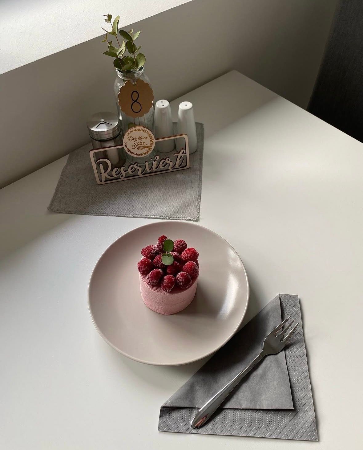 Kleines rosa Dessert mit Himbeeren auf einem Teller, daneben Besteck und eine reservierte Tischdekoration mit der Zahl 8.