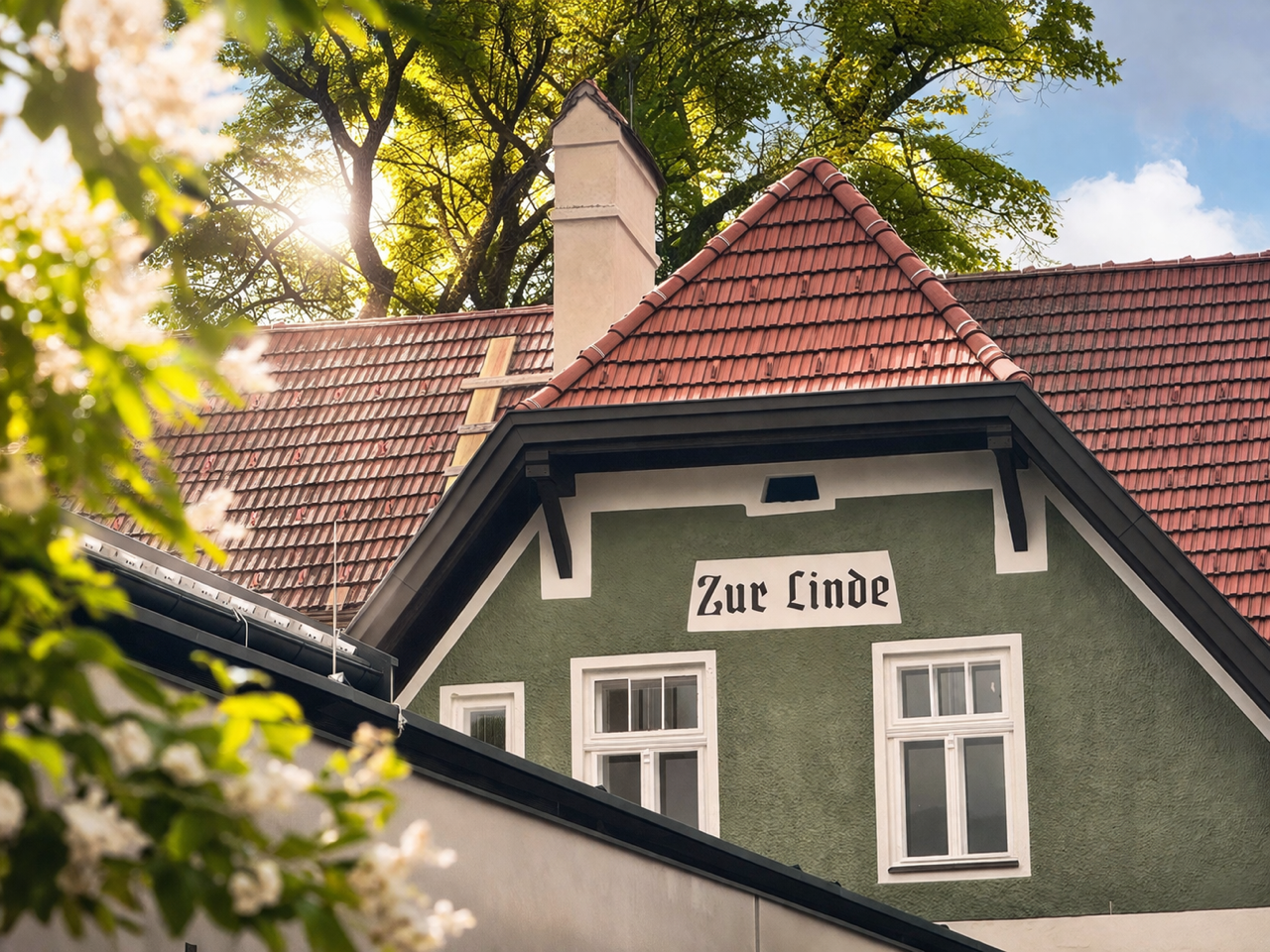 Fassade eines Gasthauses mit der Aufschrift 'Zur Linde' und roten Dachziegeln, umgeben von grünen Bäumen.