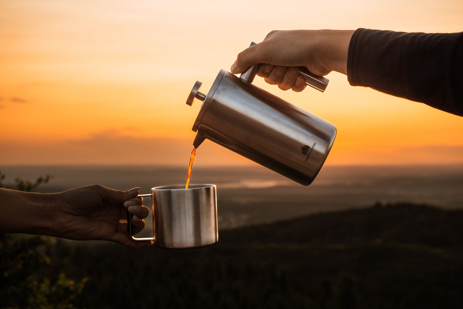 Eine Hand gießt Tee aus einer Thermoskanne in eine Tasse, vor der Kulisse eines warmen Sonnenaufgangs über bewaldeten Hügeln im Waldviertel.