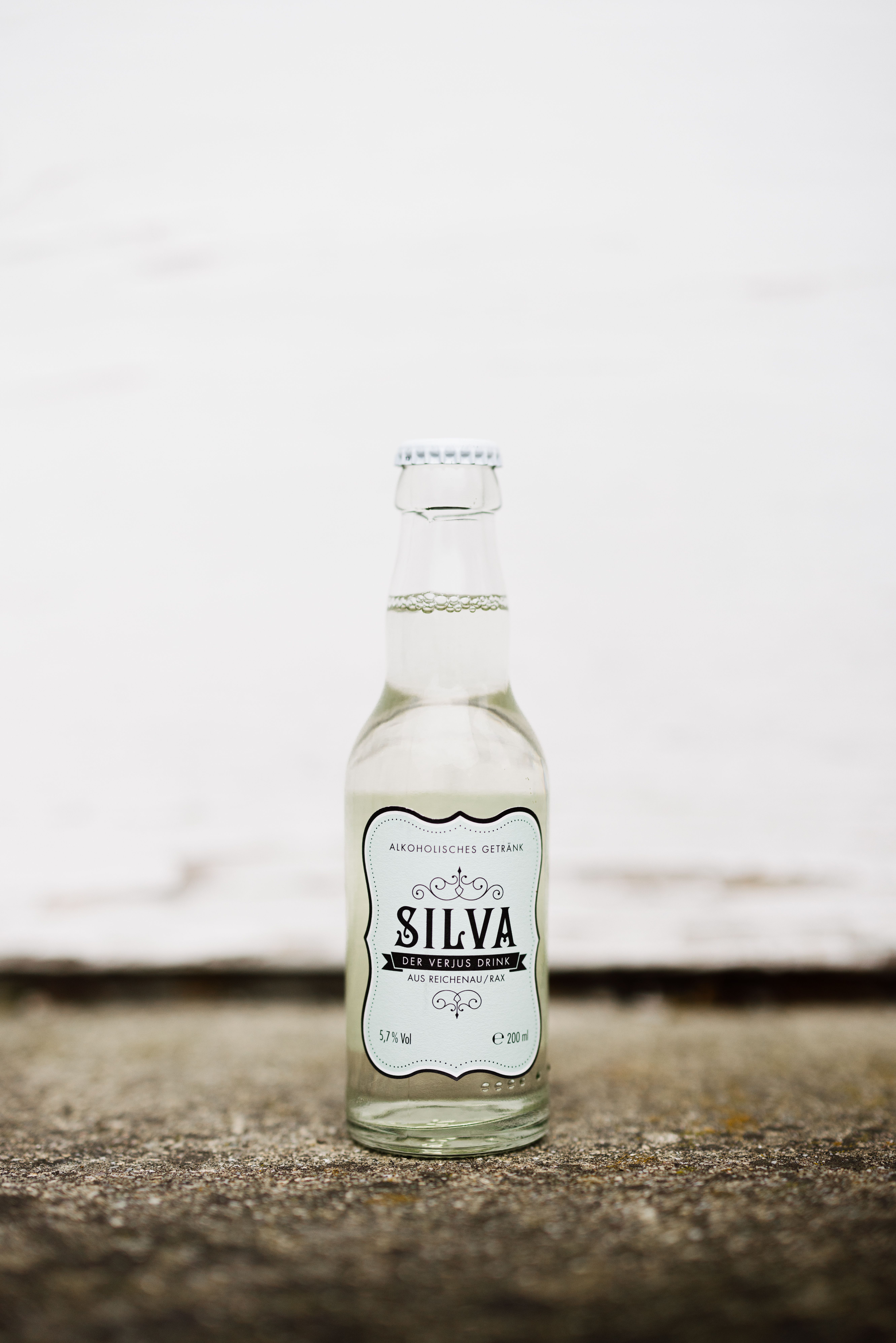 Flasche und Glas mit Silva-Verjus-Drink auf einem Betonpfeiler, unscharfe Personen im Hintergrund.