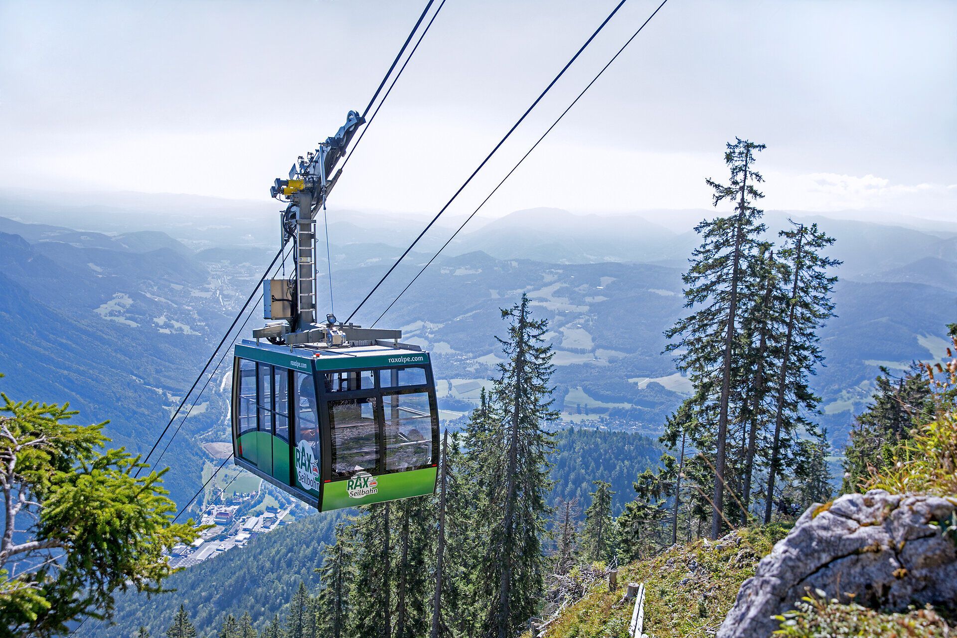 Seilbahn-Gondel über bewaldeter Berglandschaft mit weitem Blick auf Täler und Hügel bei klarem Wetter.