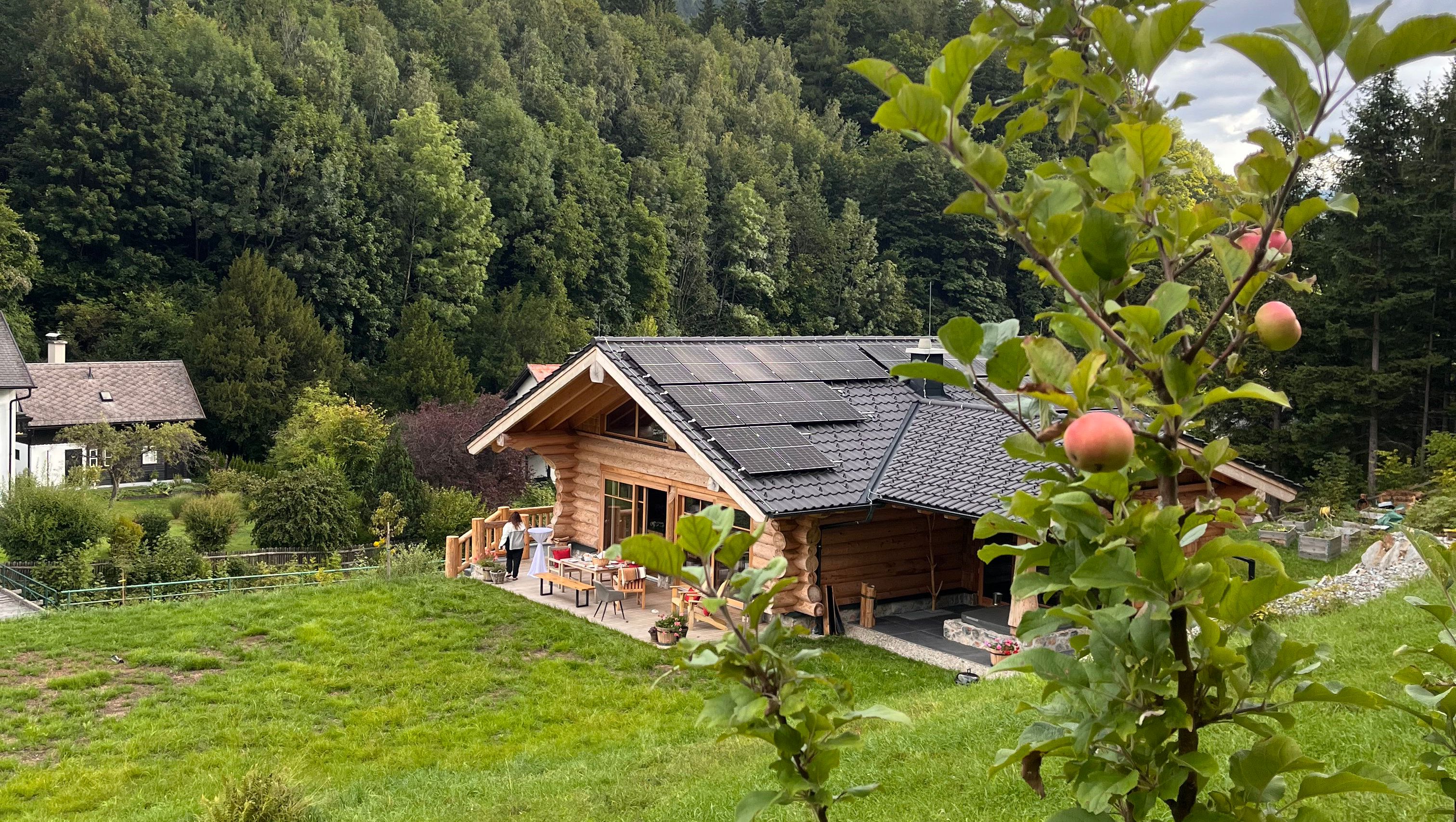 Ein Holzchalet mit Solarpanelen auf dem Dach, umgeben von grüner Landschaft und Bäumen. Im Vordergrund ein Apfelbaum mit reifen Äpfeln.