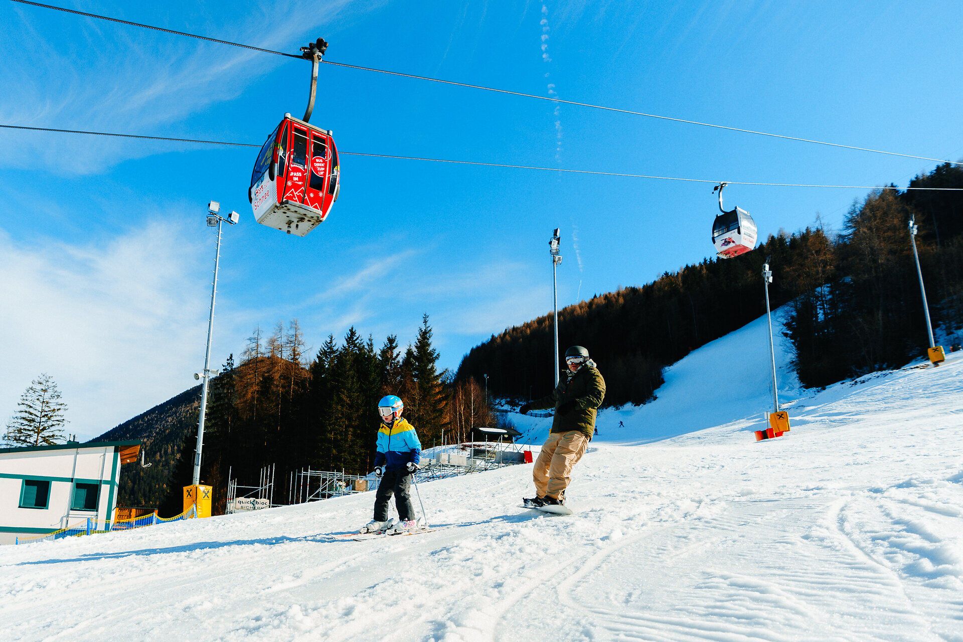 Skifahrer auf einer verschneiten Piste am Semmering Hirschenkogel, während eine rote Gondel über ihnen fährt.