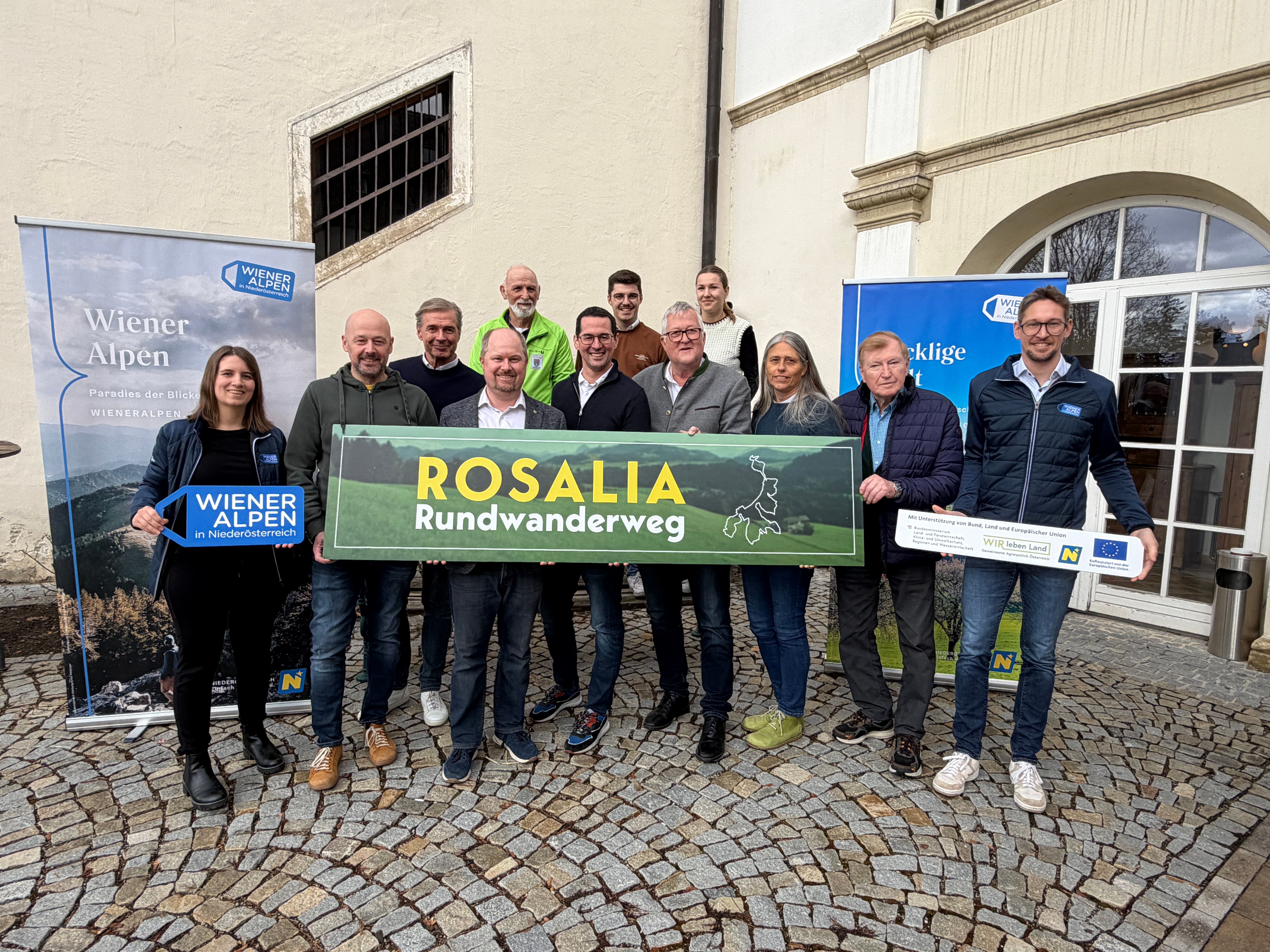 Mehrere Personen stehen in einem Innenhof und halten ein Wiener Alpen Schild, ein LEADER-Schild und ein Rosalia-Rundwanderweg-Schild in ihren Händen.