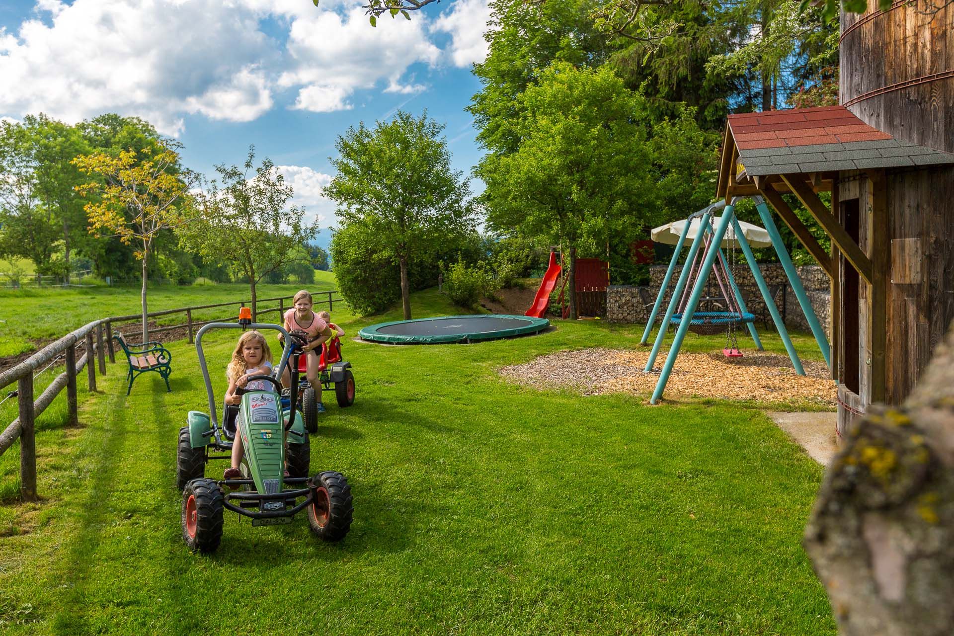 Zwei Kinder fahren auf Spielzeugtraktoren über eine Wiese. Im Hintergrund sind eine Schaukel, eine Rutsche und ein Trampolin zu sehen.