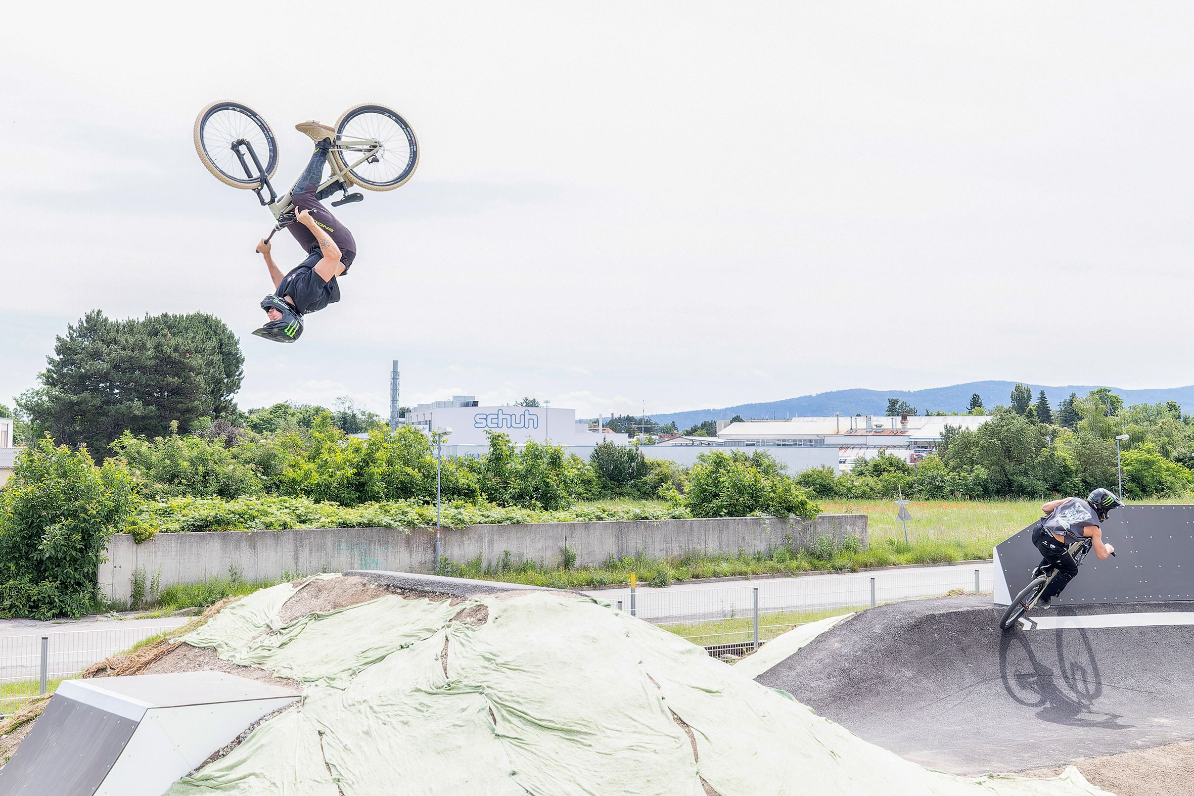 Zwei BMX-Fahrer auf einer Pumptrack-Anlage, einer macht einen Rückwärtssalto, der andere fährt eine Kurve.