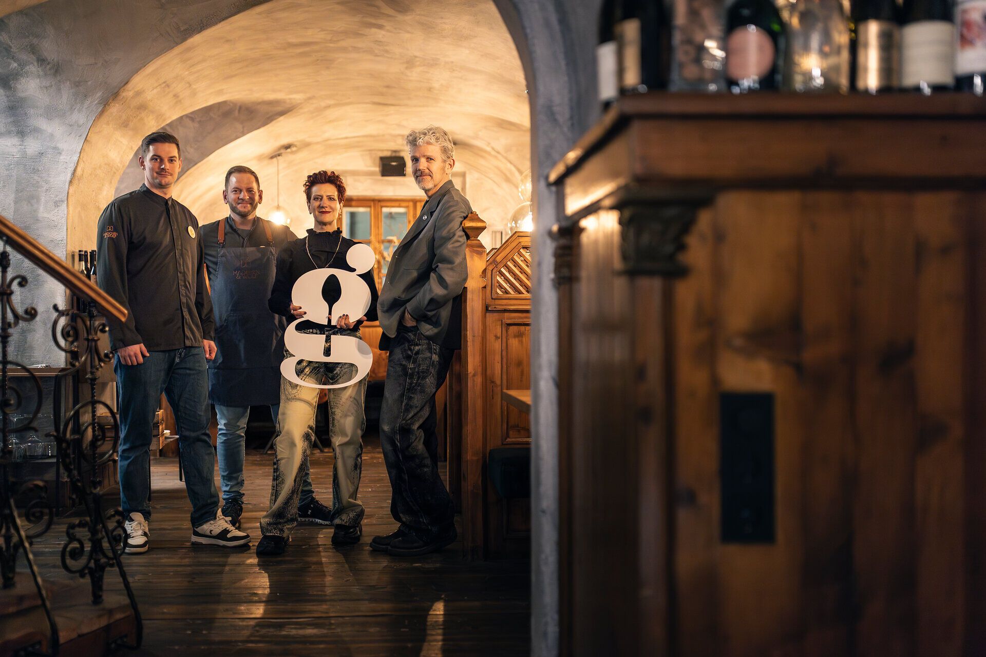 Vier Personen stehen in einer Gaststube und halten den ausgeschnittenen Buchstaben "g" in der Hand. 