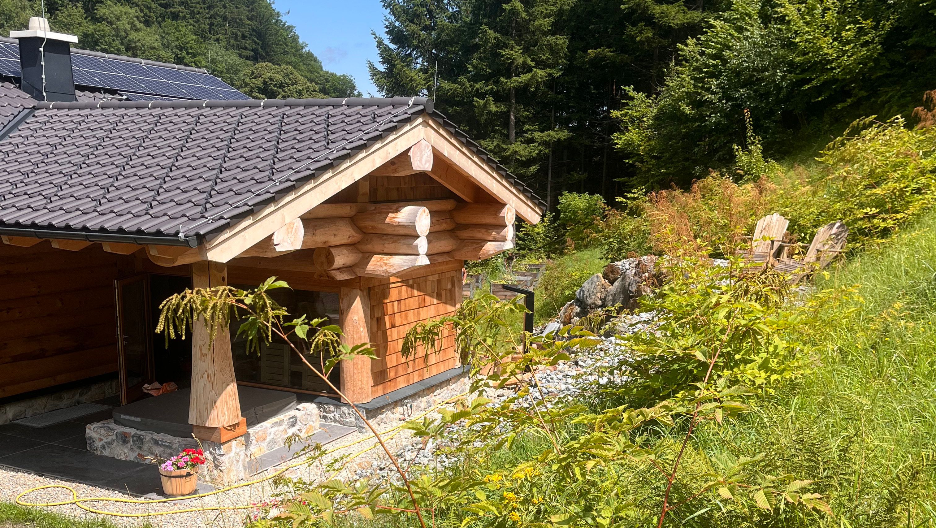 Holzchalet mit Solarpanelen auf dem Dach, umgeben von Wald und Wiese.